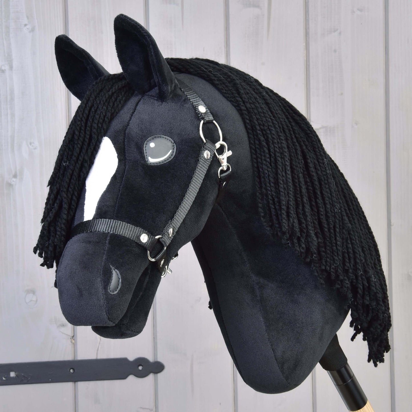 Hobby Horse Cloony avec licol noir de Hobby Horse LarDen, étalon American Quarter Horse en peluche avec tissu noir, tache blanche sur le visage, yeux en bouton, crinière noire en fil et bride sur fond en bois clair.