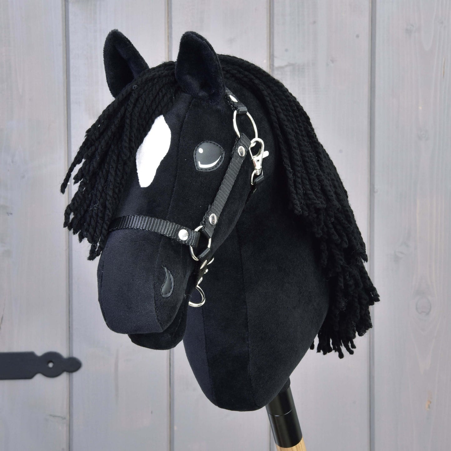 Gros plan sur Hobby Horse Cloony avec licol noir de Hobby Horse LarDen, ressemblant à un étalon American Quarter Horse, avec une tache blanche sur le front, une crinière noire en fil et un licol noir, sur fond en bois.