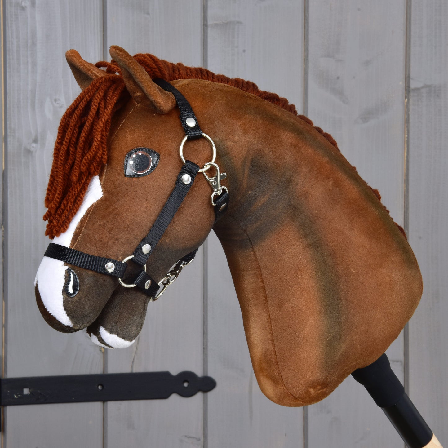 The Hobby Horse LarDen "Hobby Horse Cooper with Black Halter" features a brown plush design with a white stripe and yarn mane, set against a gray wooden background.