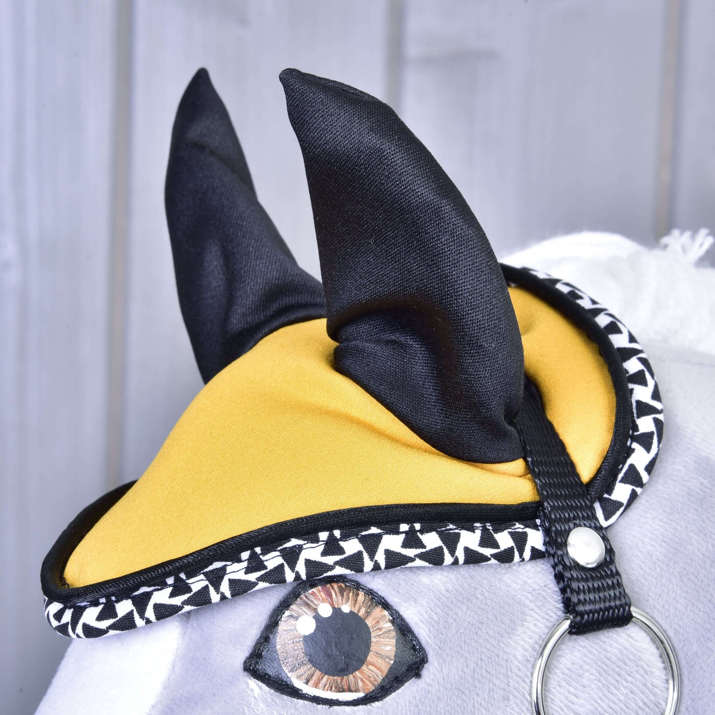 Gros plan de l’Ensemble Beauté Jaune de Hobby Horse LarDen montrant sa tête en peluche avec bonnet d’oreilles jaune et noir, bordure géométrique noire et blanche, œil peint et visage à fourrure blanche sur fond en bois gris.