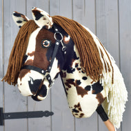 Hobby Horse Dakota mit schwarzem Halfter von Hobby Horse LarDen, ein Plüsch-Hobby Horse inspiriert von den wilden Mustangs Nordamerikas, mit braun-weißen Flecken, brauner Garnmähne und schwarzem Halfter, vor einer grauen Holzwand