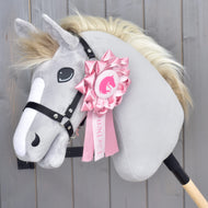 The Hobby Horse LarDen "Hobby Horse Ribbon LarDen Pink" features a gray plush horse with a black bridle and cream mane, adorned with a large pink show ribbon on its head, set against a wooden background.