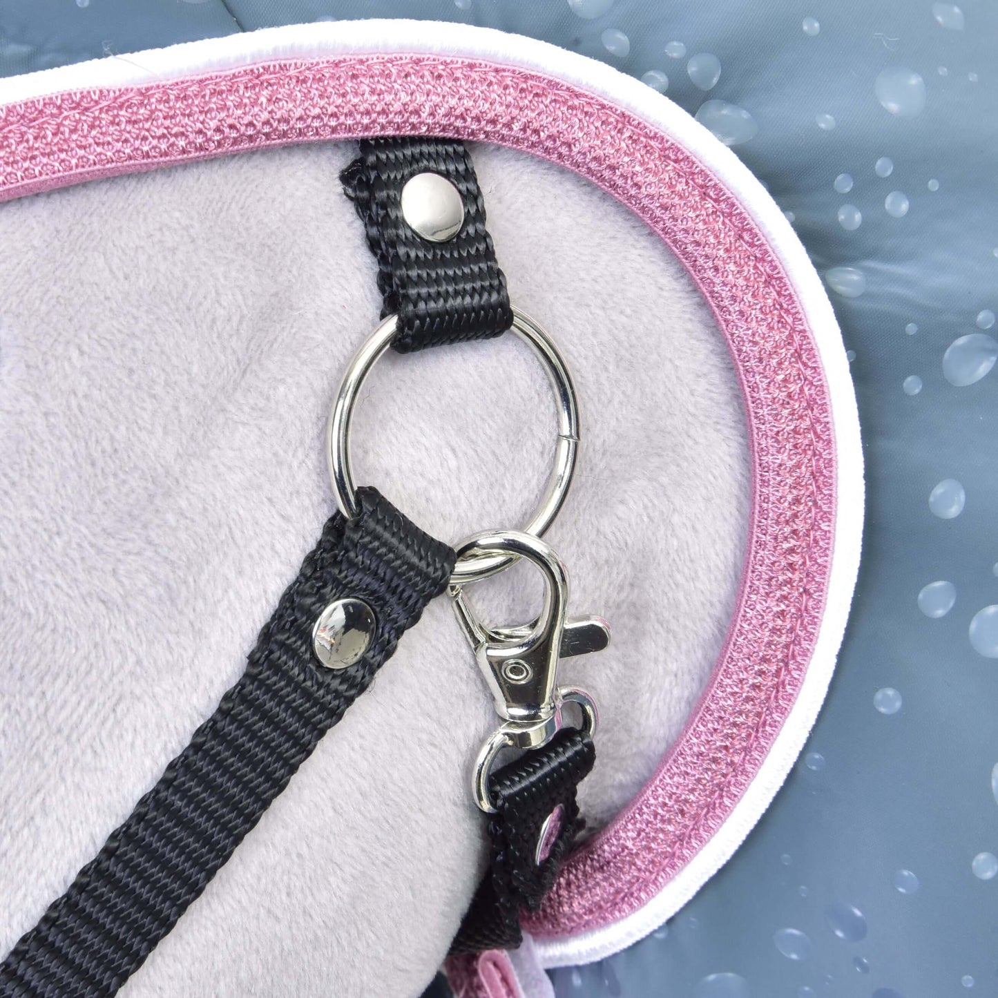 Gros plan sur une sangle noire avec mousqueton et anneau en métal attachés à l’Imperméable Best de Hobby Horse LarDen, avec tissu gris clair et bordure rose. Le fond bleu à gouttes d’eau met en valeur sa résistance par tous les temps pour le Hobby Horse.