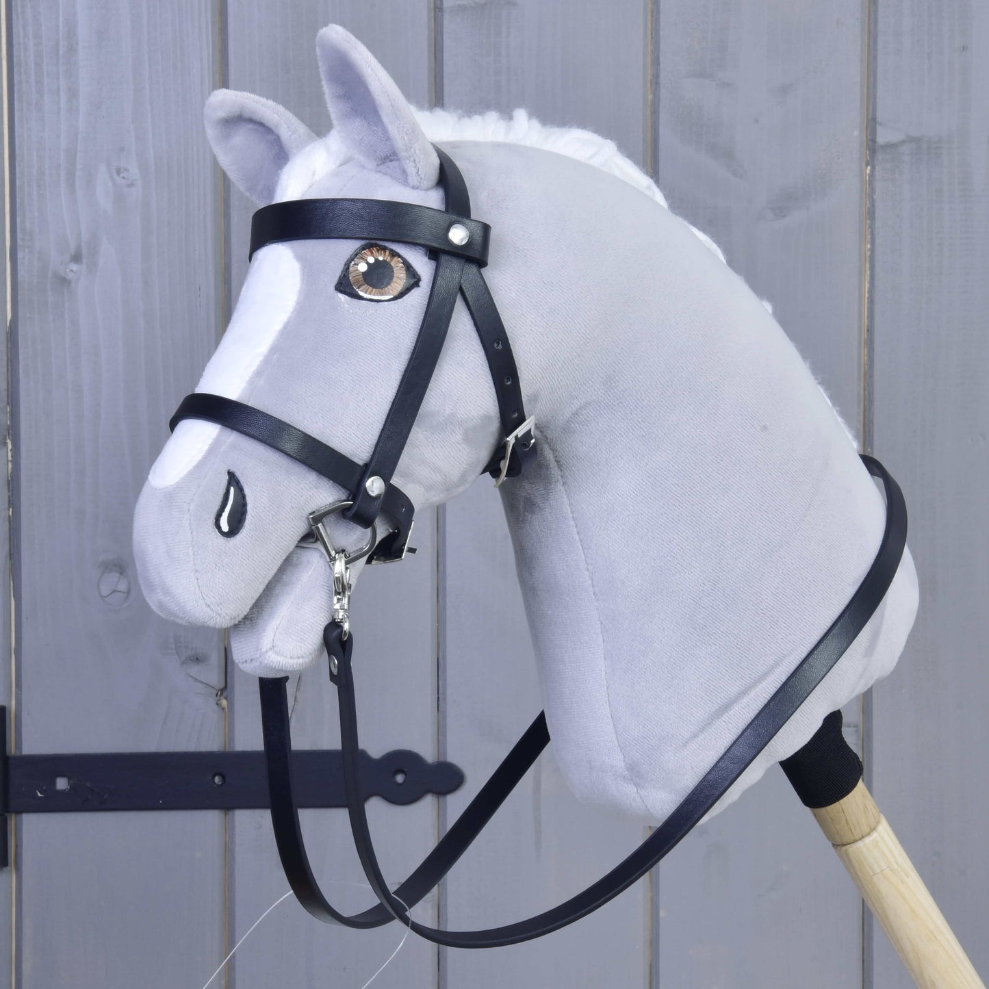 Das Hobby Horse LarDen Set Basic, ein graues Plüsch-Hobby Horse mit schwarzem Zaumzeug und Zügeln auf einem Holzstab, ist vor grauen Holzpaneelen zu sehen.