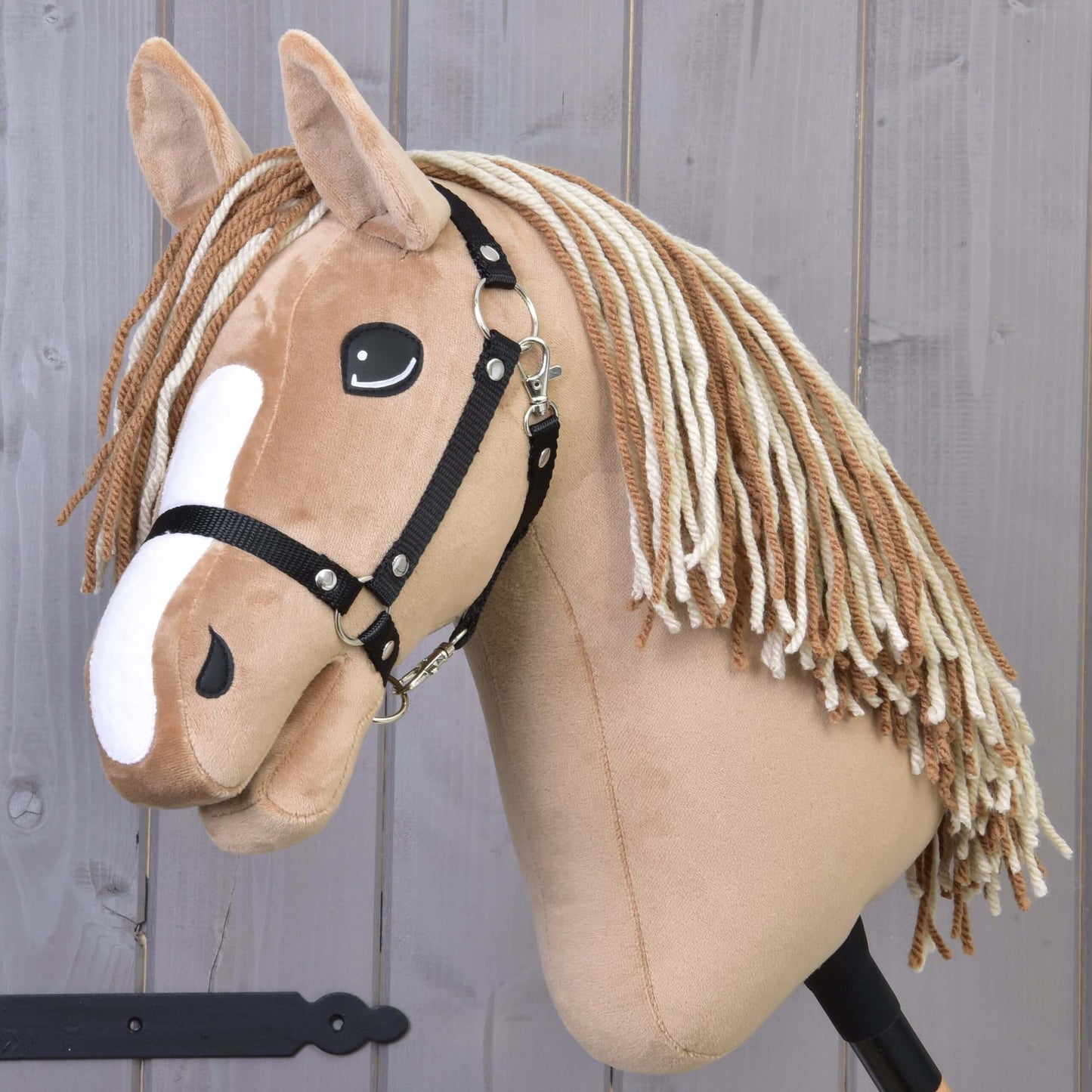 Hobby Horse Bren avec licol noir de Hobby Horse LarDen, avec corps brun clair, marques blanches sur le visage, longue crinière en fil, photographié sur fond en bois.