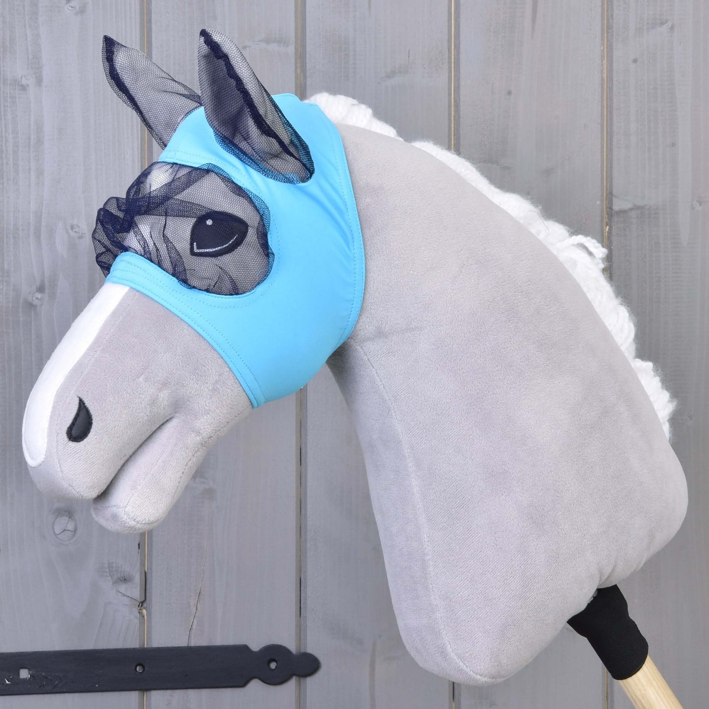 Plüsch-Hobby Horse mit hellgrauem Körper, weißer Mähne und blauer waschbarer Fliegenmaske von Hobby Horse LarDen auf einem Holzstab vor grauem Holzhintergrund.
