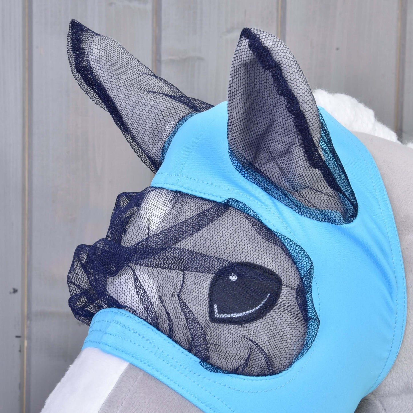 Nahaufnahme eines Plüschpferdes mit Fliegenmaske von Hobby Horse LarDen aus hellblauem Stoff mit schwarzem Mesh über Ohren, Augen und Nase zum Schutz. Die waschbare Maske ist wie bei echten Pferden gestaltet und vor einem Holzhintergrund gezeigt.