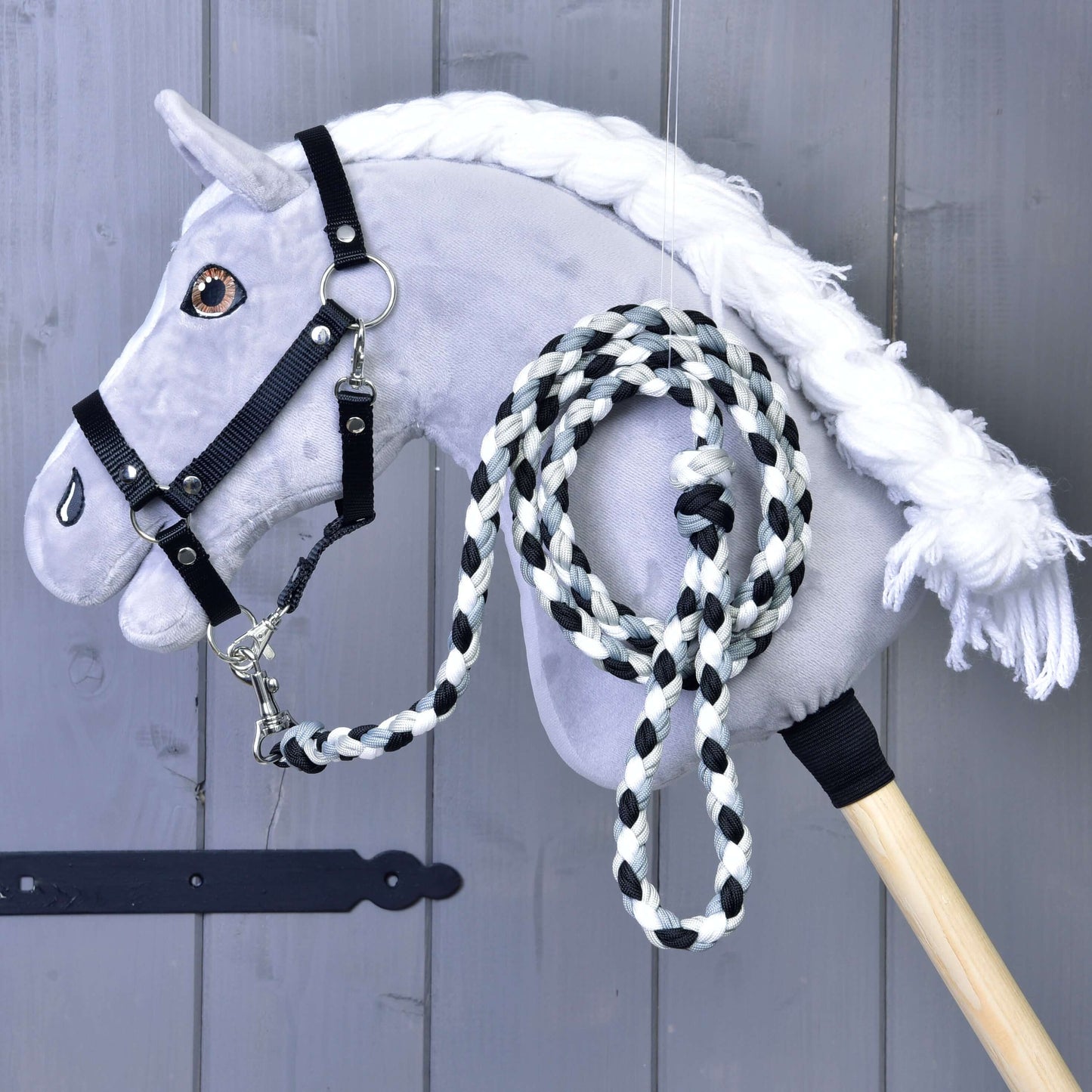 Hobby Horse - gestrickter Führstrick schwarz/weiß von Hobby Horse LarDen mit grauem Plüschkopf, weißer Mähne, schwarzem Zaumzeug und inklusive schwarz-weiß geflochtenem Führstrick, präsentiert vor einem grauen Holzhintergrund.