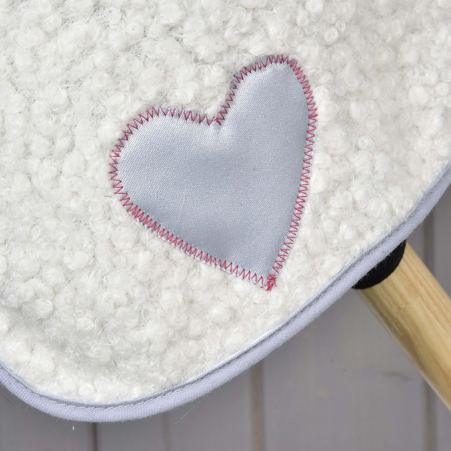Nahaufnahme der weißen strukturierten Fliegenhaube Set Snowflake von Hobby Horse LarDen mit glänzendem hellgrauem Herz-Aufnäher und rosa Zickzackstichen; unten rechts ist ein Holzstab zu sehen.