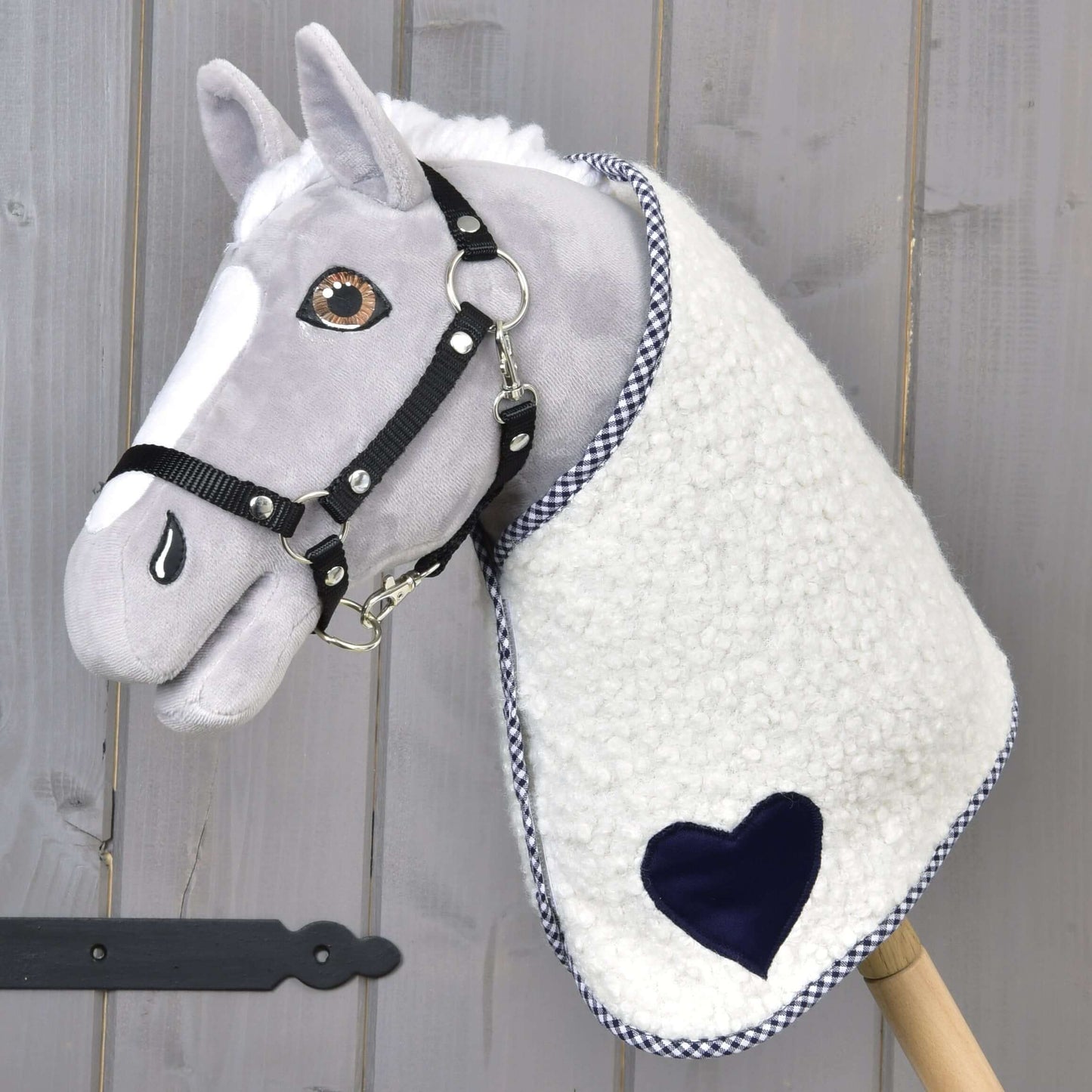 Decke Cube Blue von Hobby Horse LarDen ist ein plüschiges Hobby Horse mit grauem Gesicht, schwarzem Zaumzeug und einer Decke mit blauen Akzenten, auf einem Holzstab vor Holz-Hintergrund.