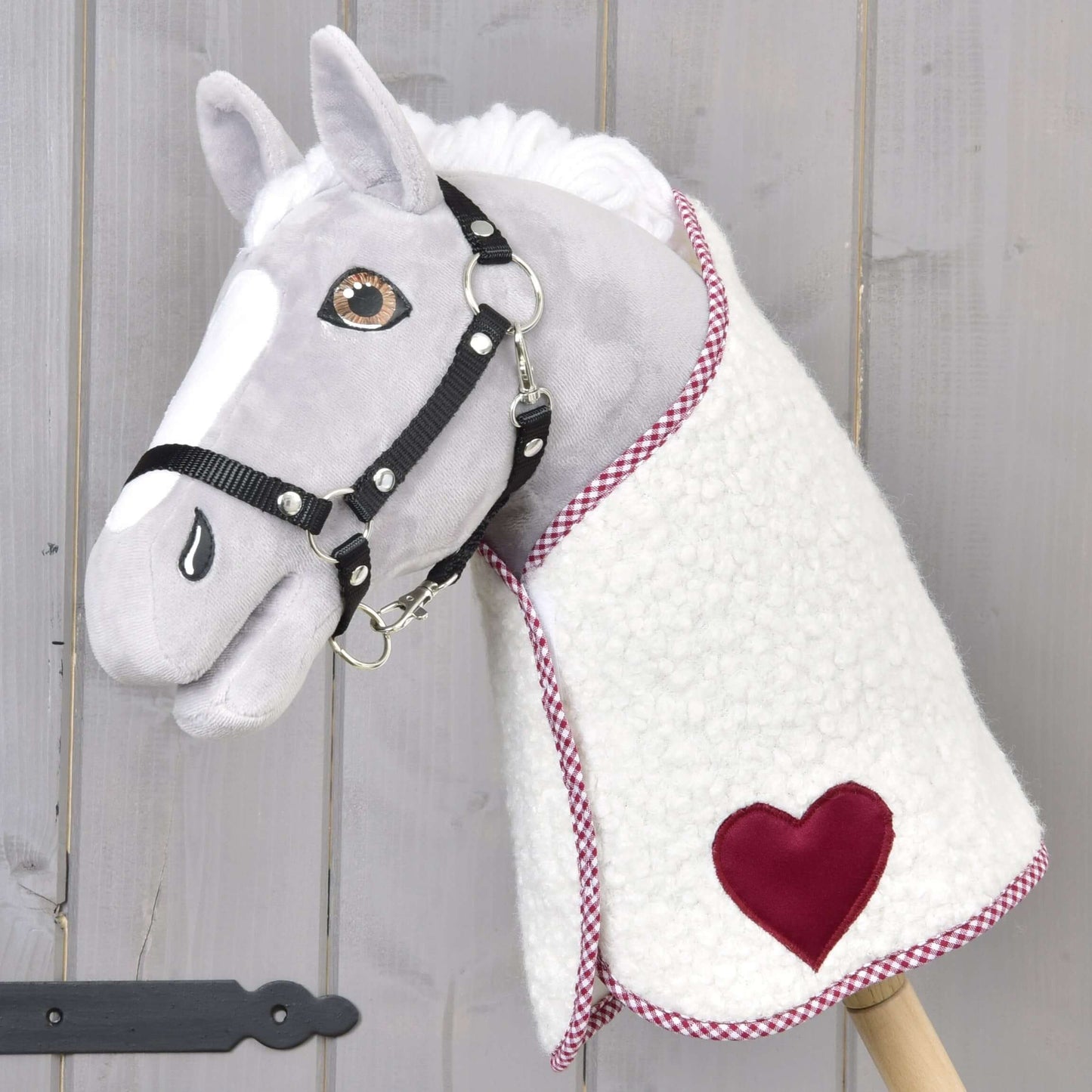Das plüschige Hobby Horse von Hobby Horse LarDen hat ein weiß-graues Gesicht, ein schwarzes Zaumzeug und trägt die Decke Cube Red aus Fleece mit rotem Herzaufnäher, montiert auf einem Holzstab vor hellem Holz.
