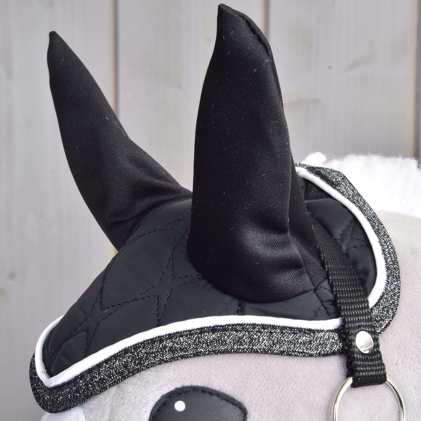 Nahaufnahme des Set Thermo Black Queen von Hobby Horse LarDen mit plüschigen schwarzen Ohren mit Kappe, einer gesteppten Abdeckung mit Glitzereinfassung wie eine Hobby Horse-Decke und einem Riemen mit Metallring vor heller Holzvertäfelung.