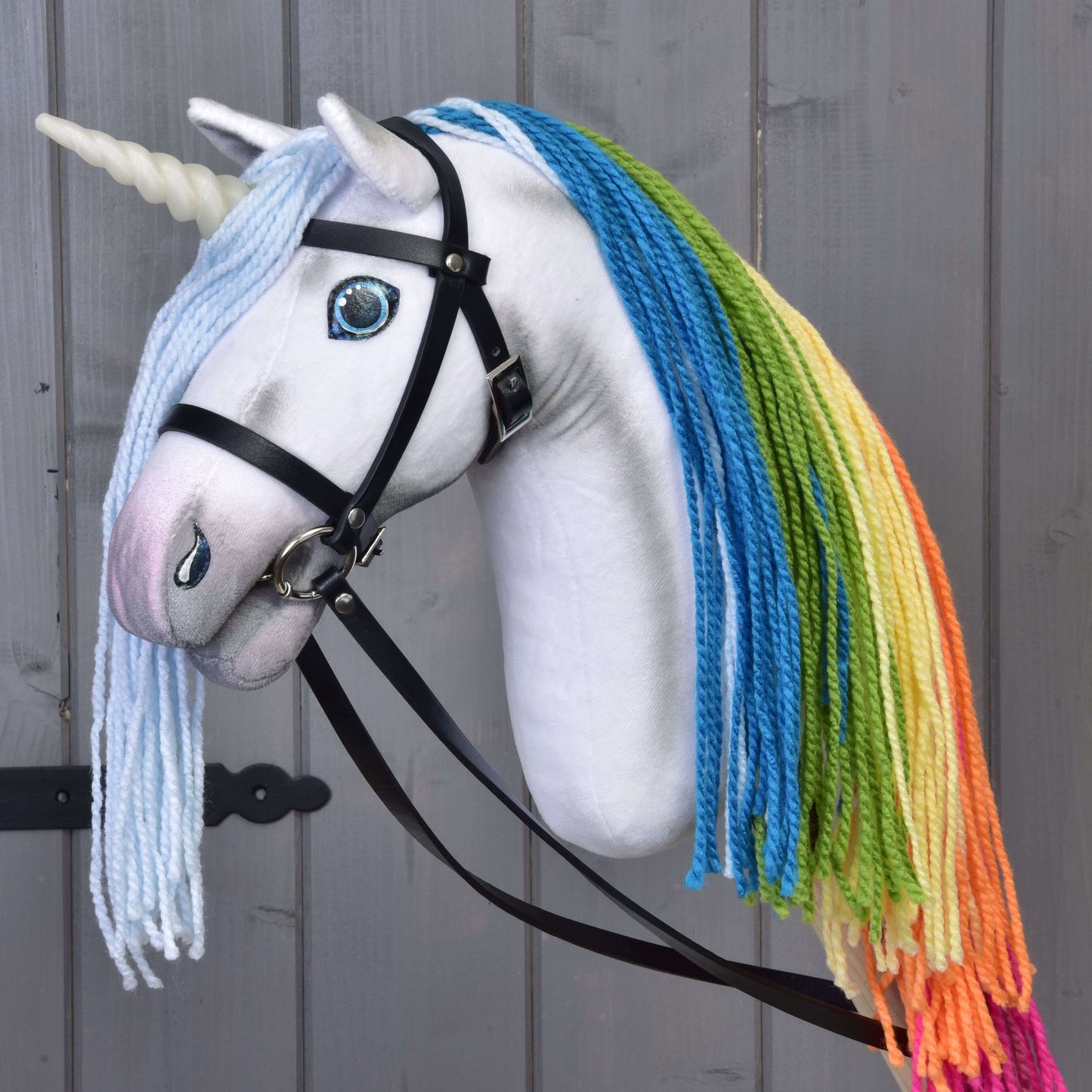 Hobby Horse LarDen Hobby Horse Einhorn Rainbow mit Trense und Halfter mit weißem Horn, blauen Augen, schwarzem Zaumzeug und bunter Regenbogenmähne aus Garn, vor grauem Holzhintergrund.