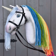 The Hobby Horse LarDen Hobby Horse Unicorn Rainbow with Bridle and Halter features a white horn, blue eyes, black bridle, and colorful rainbow yarn mane, shown against a gray wooden background.