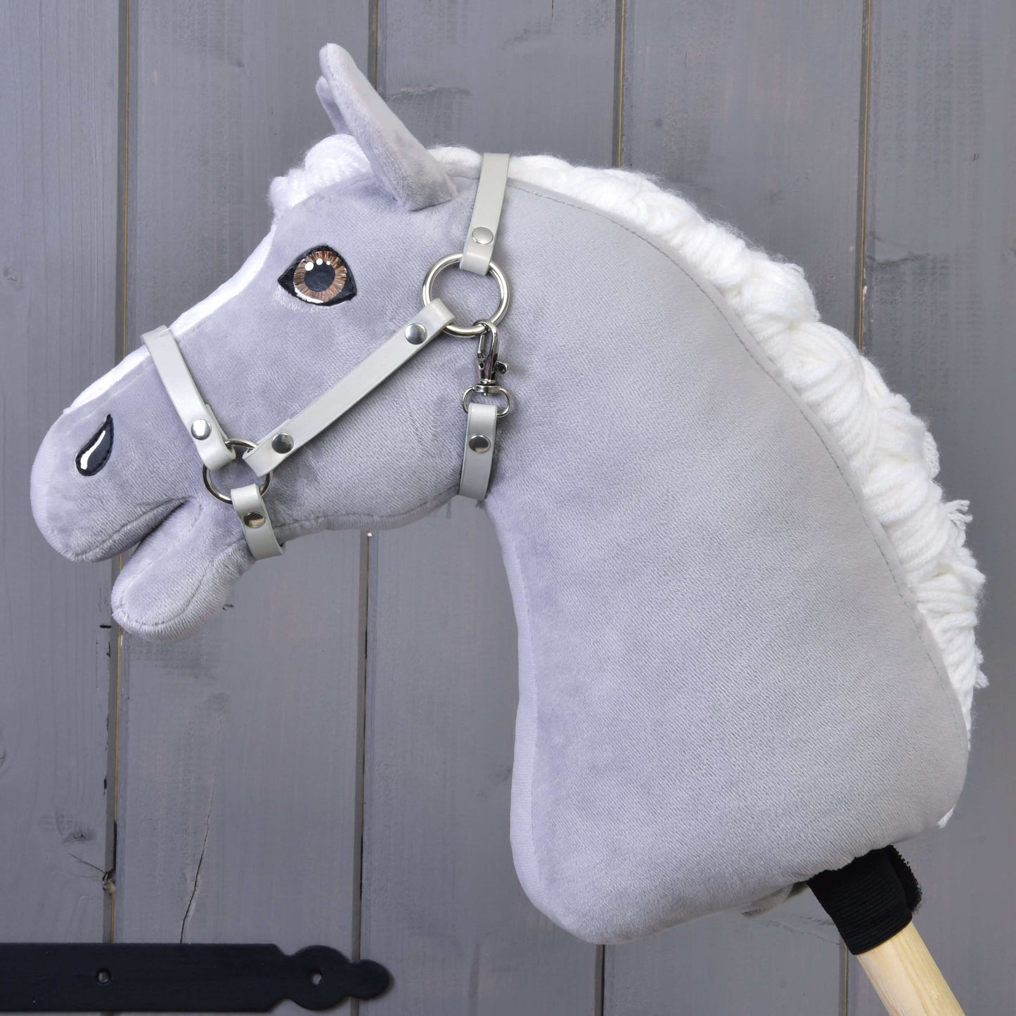 Licol - similicuir gris clair (taille S) de Hobby Horse LarDen, présenté sur un cheval en peluche gris avec crinière blanche, yeux marron, monté sur un bâton en bois et montré devant un fond en bois gris.