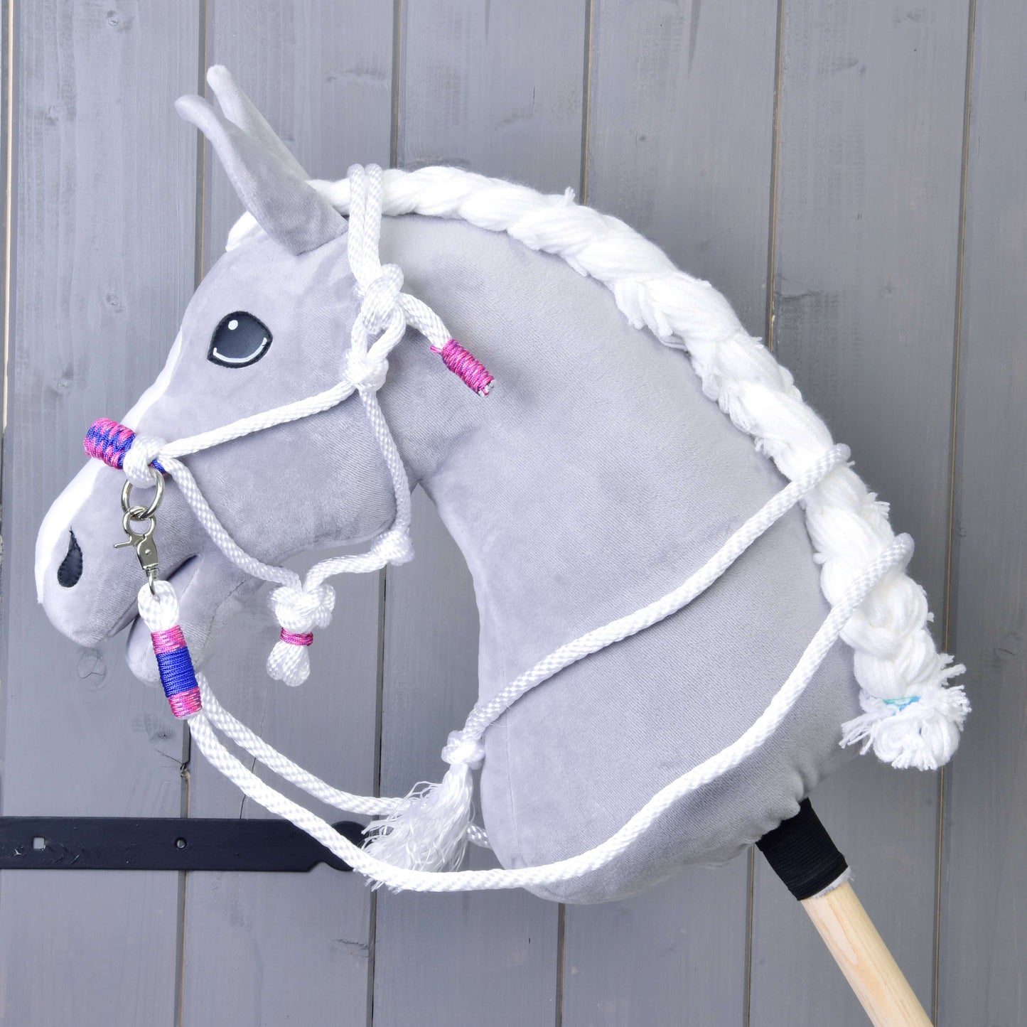 Seilset Weiß (Gr. M) von Hobby Horse LarDen an einem grauen Plüsch-Hobby Horse mit weißer geflochtener Mähne und rosa, blauen und weißen Zügeln vor grauem Holzhintergrund.