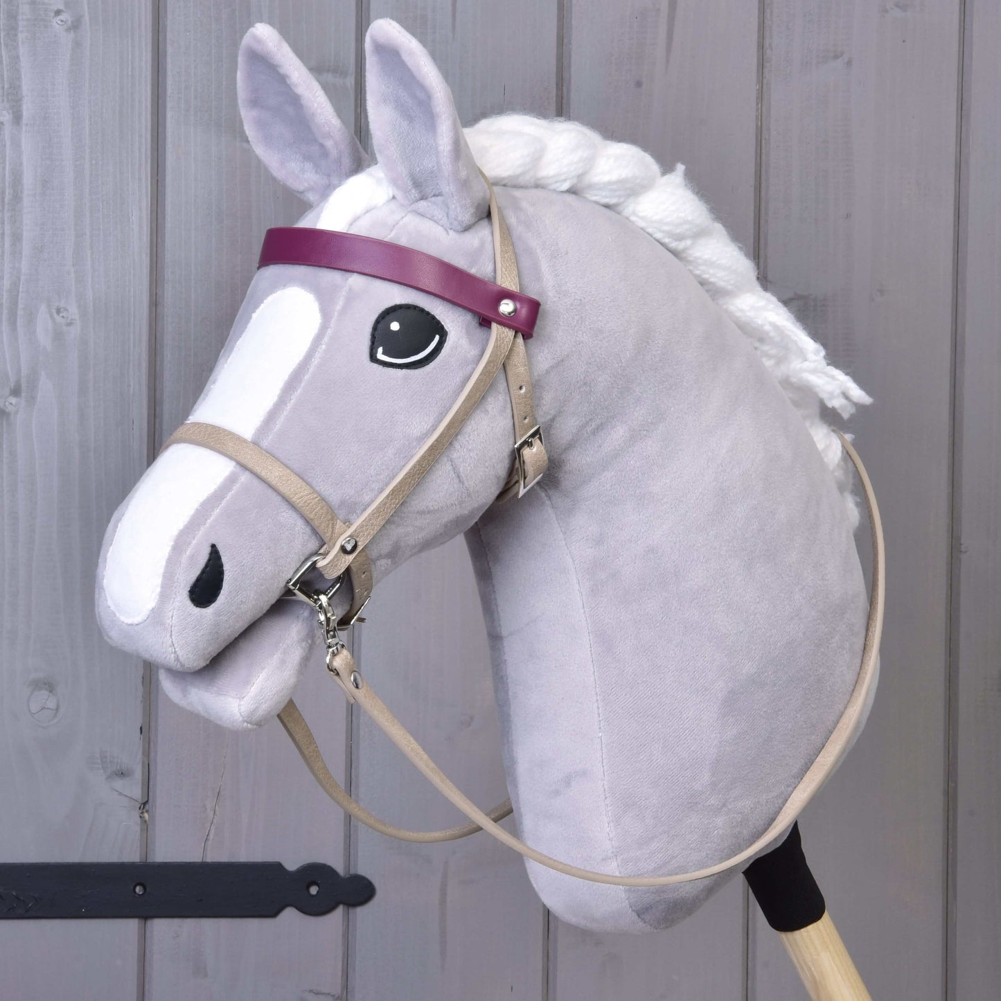 The Hobby Horse LarDen features a gray hobby horse with a white mane, black eye, and a Bordeaux leatherette set—bridle, reins, and martingale—plus a purple browband and cartoon-like face. It stands upright against a wooden background.