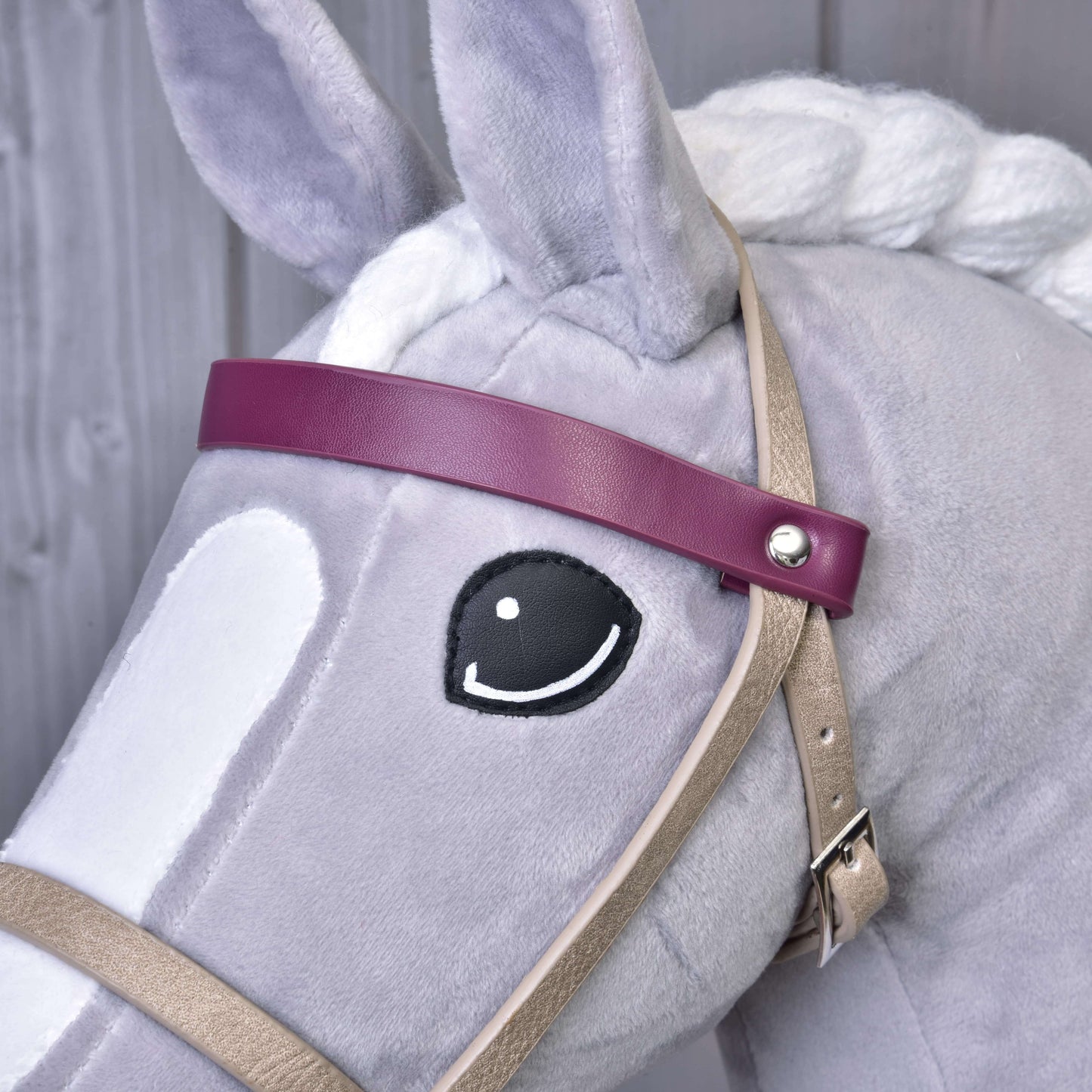 A close-up of a Hobby Horse LarDen with a white mane and stitched black eye, wearing the Bordeaux leatherette set—bridle, reins, and martingale. It sports a purple headband and is pictured against a wooden background.