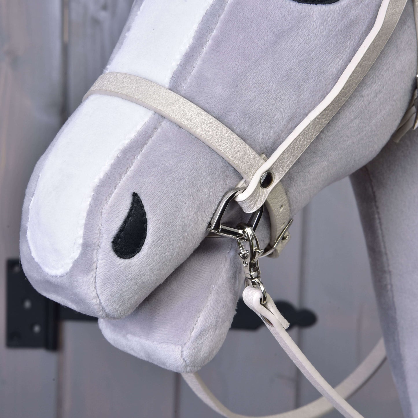 Gros plan sur la tête d’un Hobby Horse gris et blanc portant l’ensemble similicuir Aqua - bride, rênes et martingale de Hobby Horse LarDen. La bride a des ferrures métalliques. L’arrière-plan flou montre une surface en bois et une charnière métallique sombre.