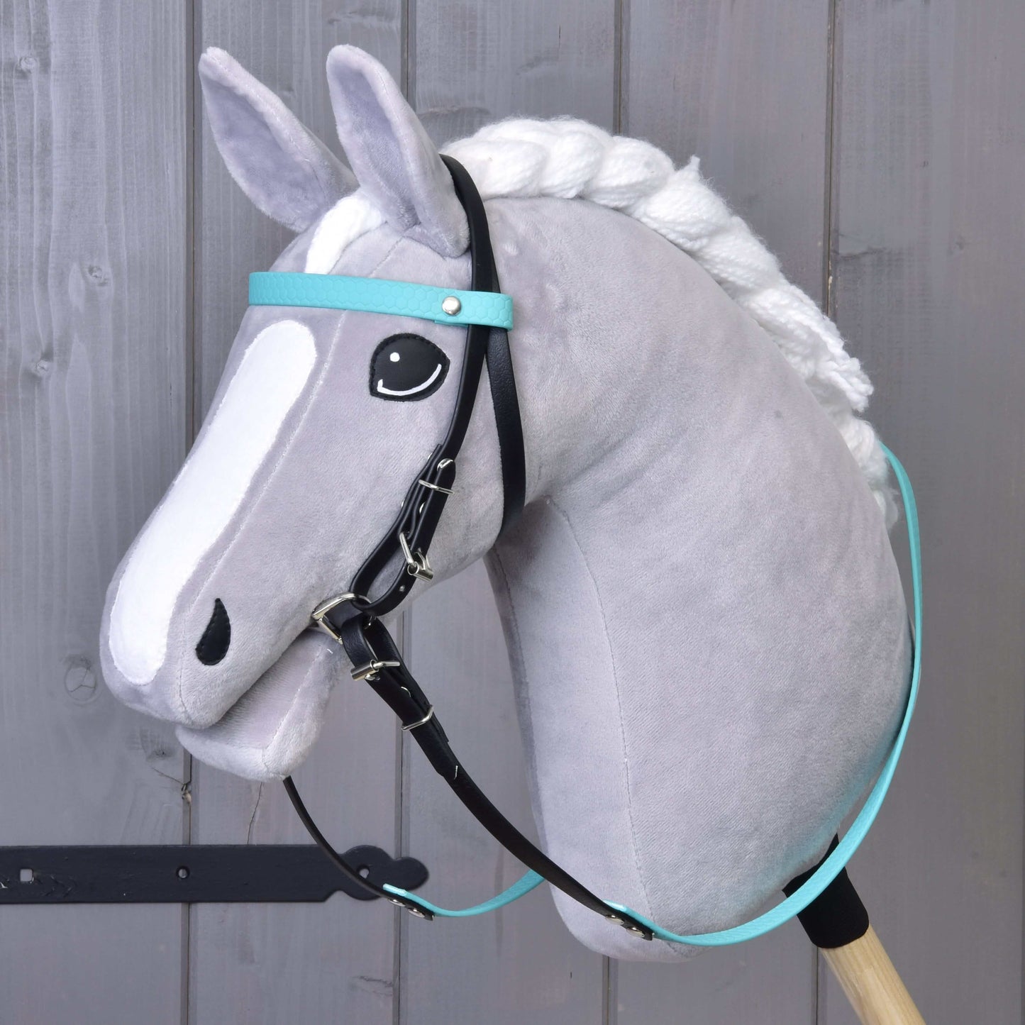 Ein graues Plüsch-Hobby Horse mit weißer Mähne und Gesichtsabzeichen trägt das Hobby Horse LarDen Komplettes Zaumzeug ohne Nasenriemen Türkis, schwarze Zügel und ist auf einem Holzstab vor einer grauen Holzwand montiert.