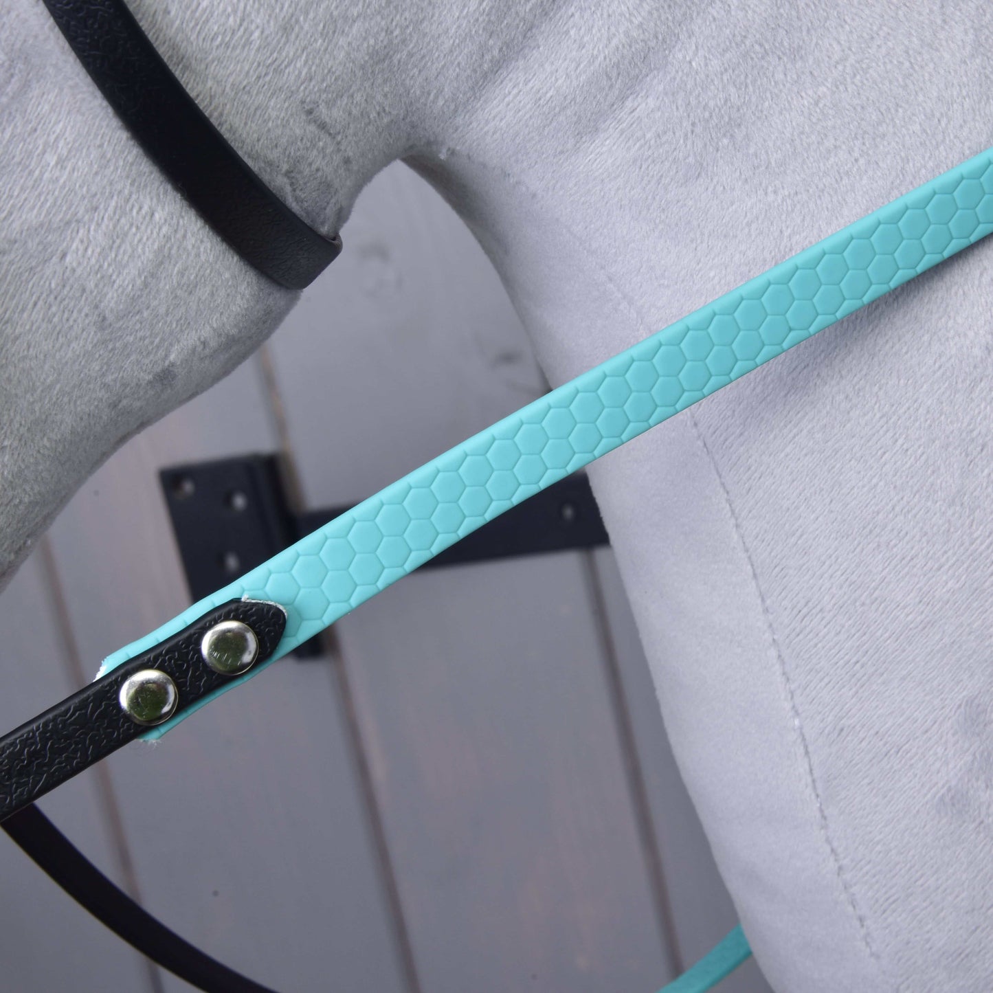 Nahaufnahme des Hobby Horse LarDen Komplettes Zaumzeug ohne Nasenriemen Türkis mit einem strukturierten Kunstlederriemen in Wabenoptik mit Metallnieten, befestigt an einem schwarzen Riemen auf einer hellgrauen Oberfläche vor unscharfem Holz-Hintergrund.