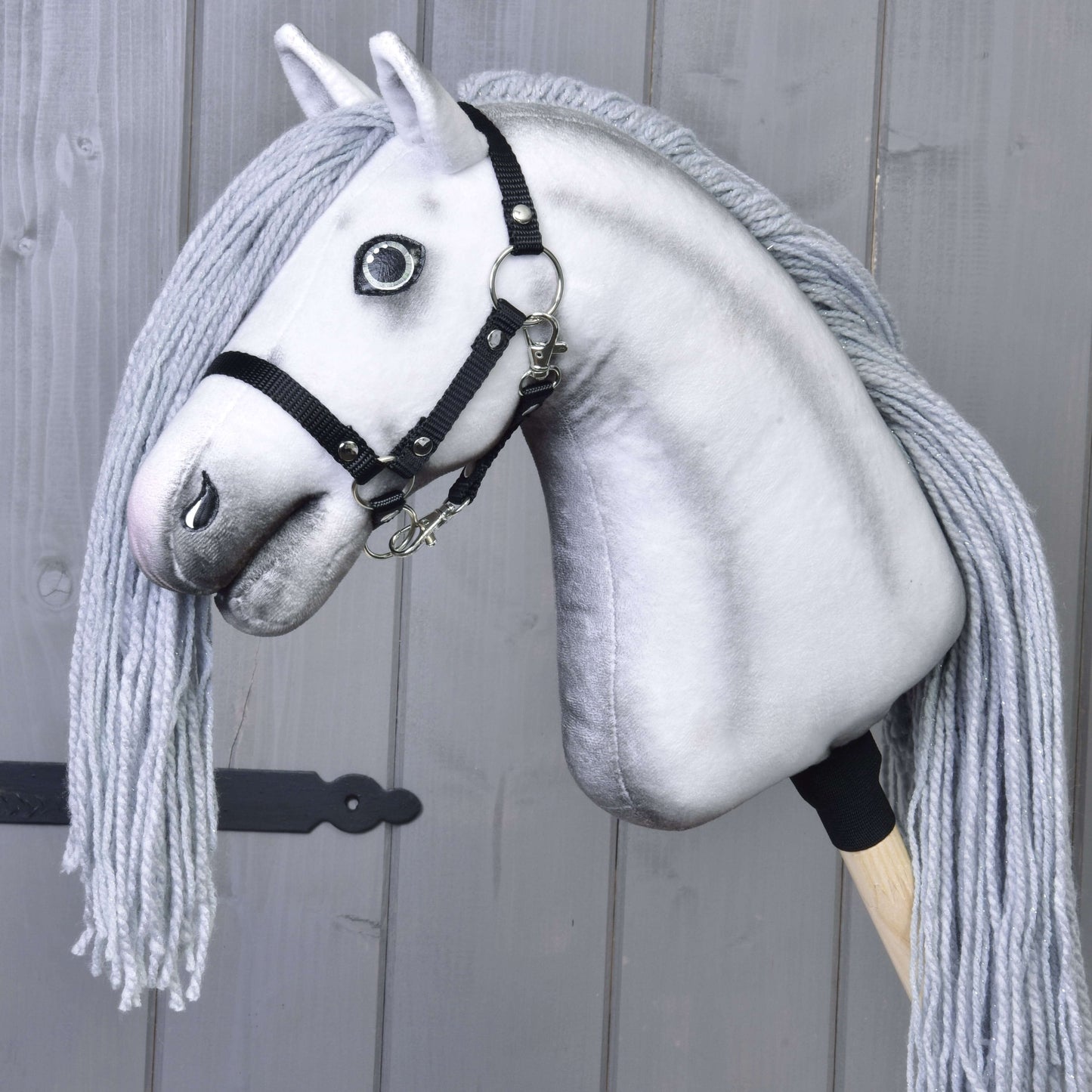 Hobby Horse Einhorn Silver mit Trense und Halfter von Hobby Horse LarDen mit schwarzer Trense, Halfter und langer Garnmähne, einem magischen Einhorn nachempfunden, vor grauer Holzwand.