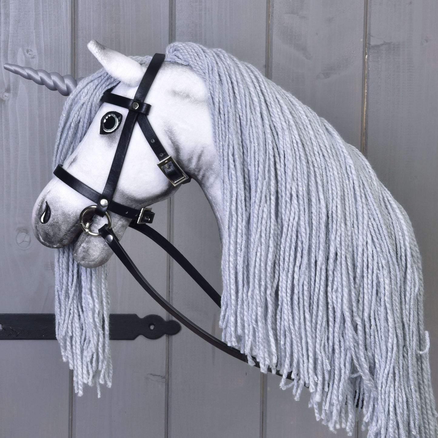 Hobby Horse Einhorn Silver mit Trense und Halfter von Hobby Horse LarDen mit weißem Gesicht, grauer Garnmähne, hellgrauem Horn und schwarzer Trense, auf einem Stock montiert und vor Holz-Hintergrund fotografiert.