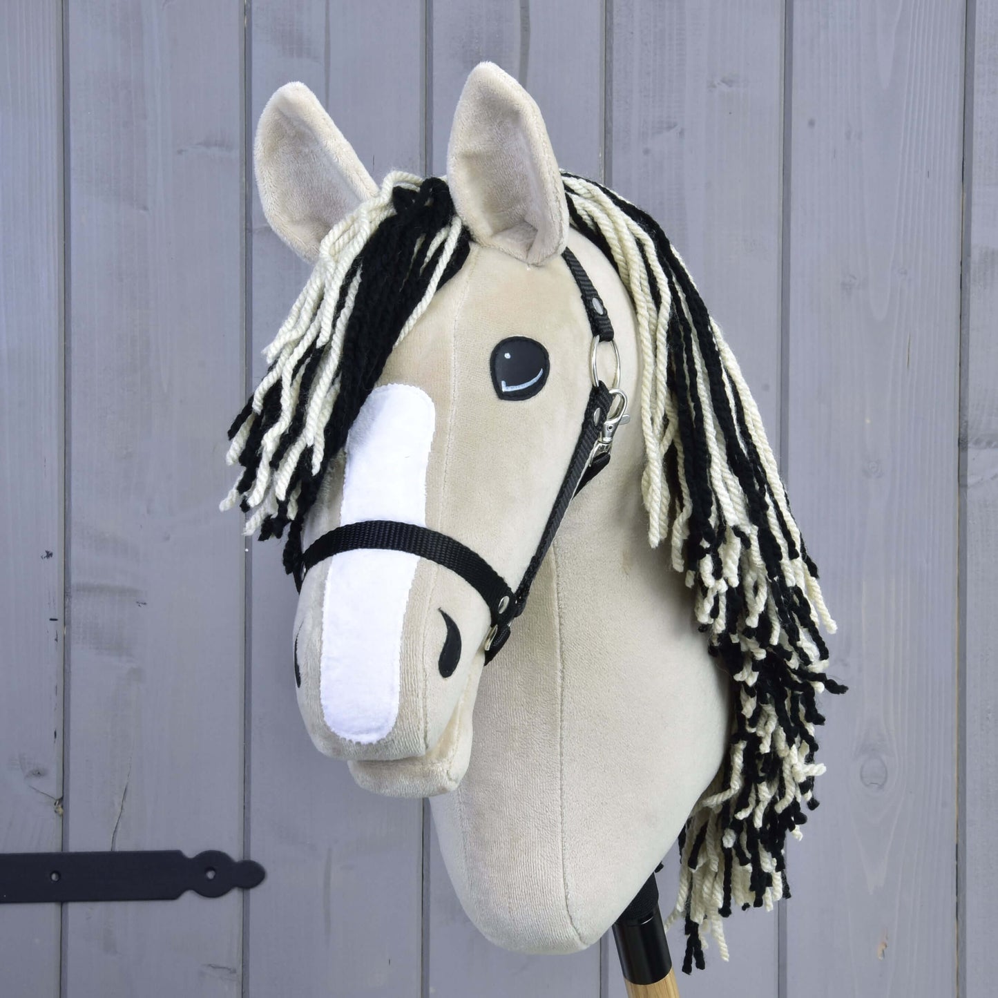 Hobby Horse Aurora mit Schwarzem Halfter von Hobby Horse LarDen mit beigem Plüschdesign, inspiriert vom American Quarter Horse, schwarzem Halfter, schwarz-weißer Garnmähne, weißem Nasenstreifen aus Filz und auf einem Holzstab vor einer hellgrauen Holzwand.