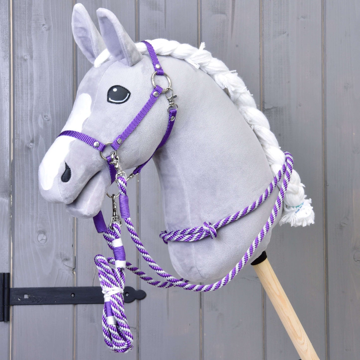 Set Start Lila (Halfter + Halsring + Führstrick + Zügel) von Hobby Horse LarDen an einem Plüsch-Hobby Horse mit grauem Kopf, weißer Mähne und weißer Blesse, auf einem Holzstab montiert vor einer grauen Holzwand.