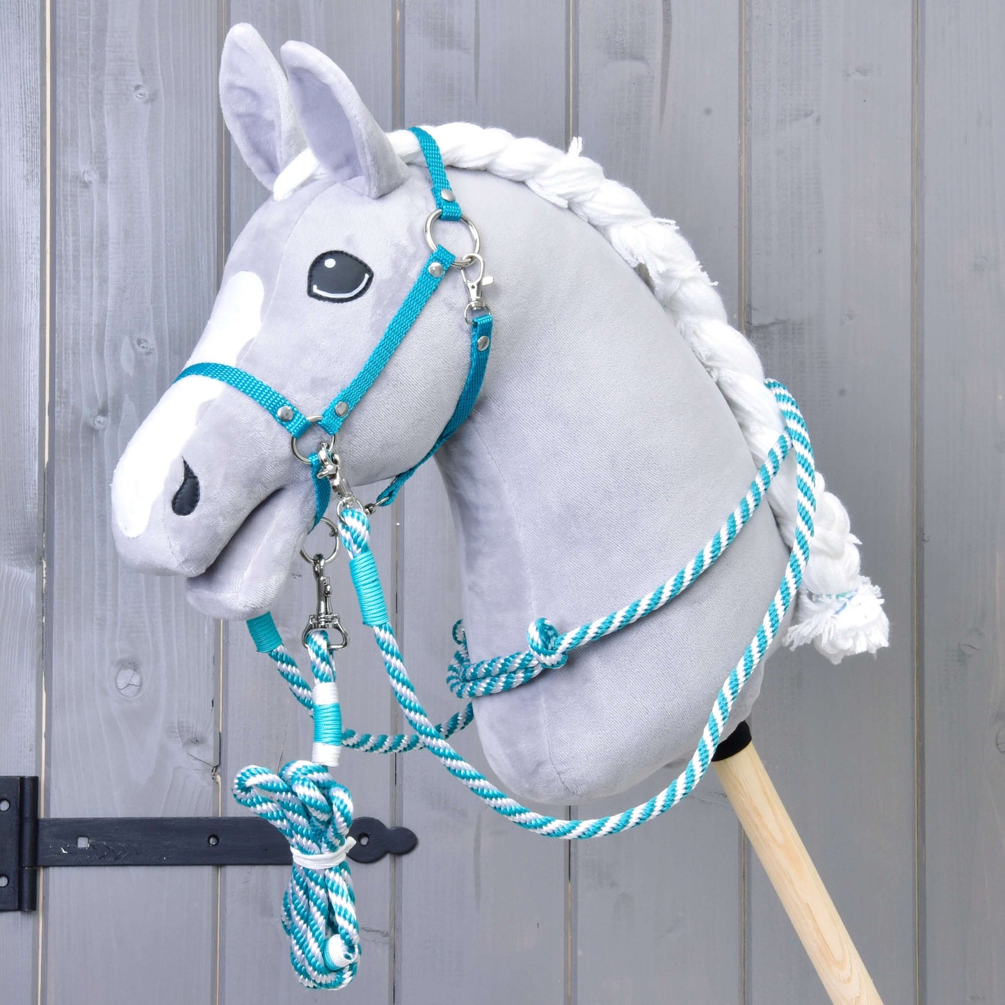 Ein graues Plüsch-Hobby Horse mit weißer Mähne ist mit dem Set Start Turqouise (halter + cordeo + lead rope + reins) von Hobby Horse LarDen ausgestattet und wird vor grauem Holzhintergrund gezeigt.