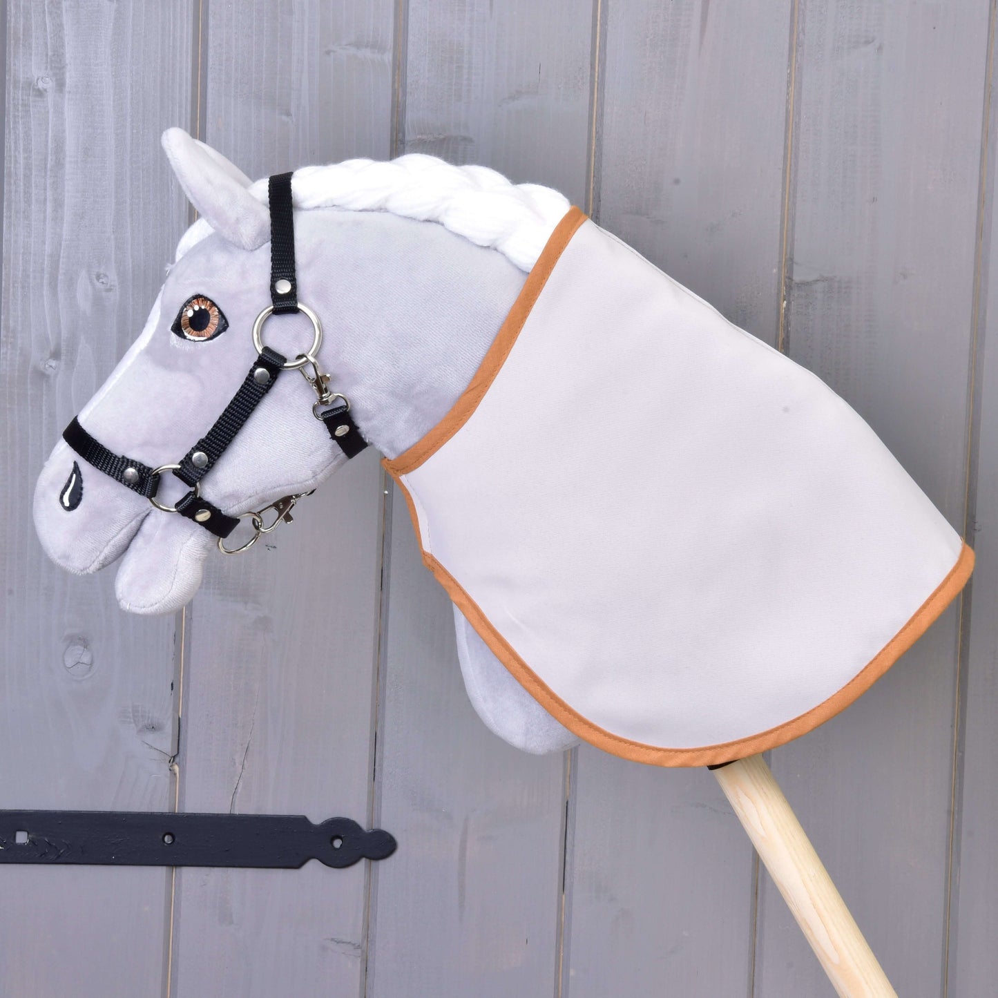 Graues Hobby Horse LarDen mit weißer Mähne, schwarzem Zaumzeug und Decke englischer Karostoff, braun & doppelseitig auf einem Holzstab vor einer grauen Holzwand.
