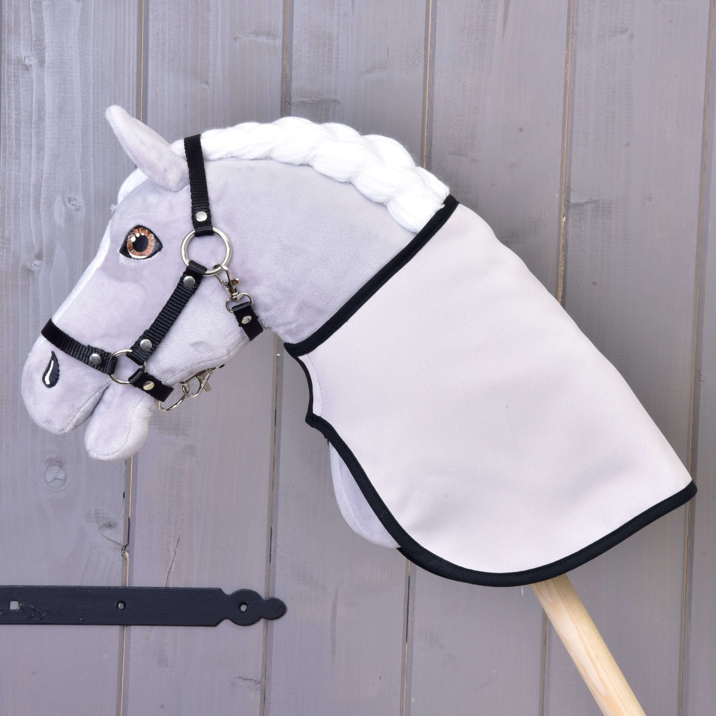 Graues Plüsch-Hobby Horse mit weißer Mähne, schwarzem Zaumzeug und Decke englischer Karostoff, schwarz & doppelseitig von Hobby Horse LarDen, auf einem Holzstab vor einer Holzwand aus Planken.