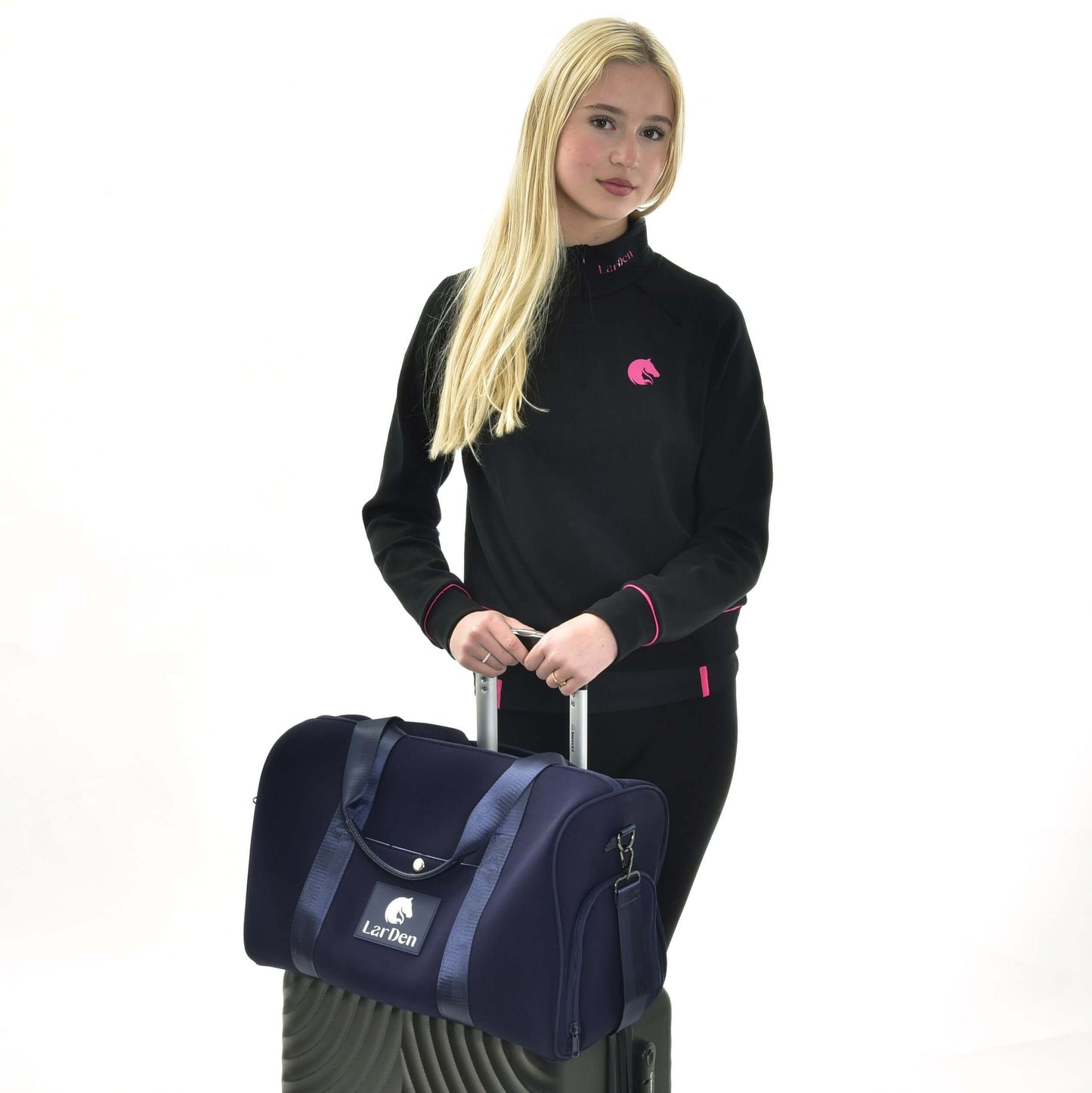 Personne aux longs cheveux blonds, portant un sweat noir avec détails roses, tenant la sangle réglable du Sac de sport LarDen bleu marine de Hobby Horse LarDen, placé sur une valise noire sur fond blanc. Idéal pour un usage quotidien.