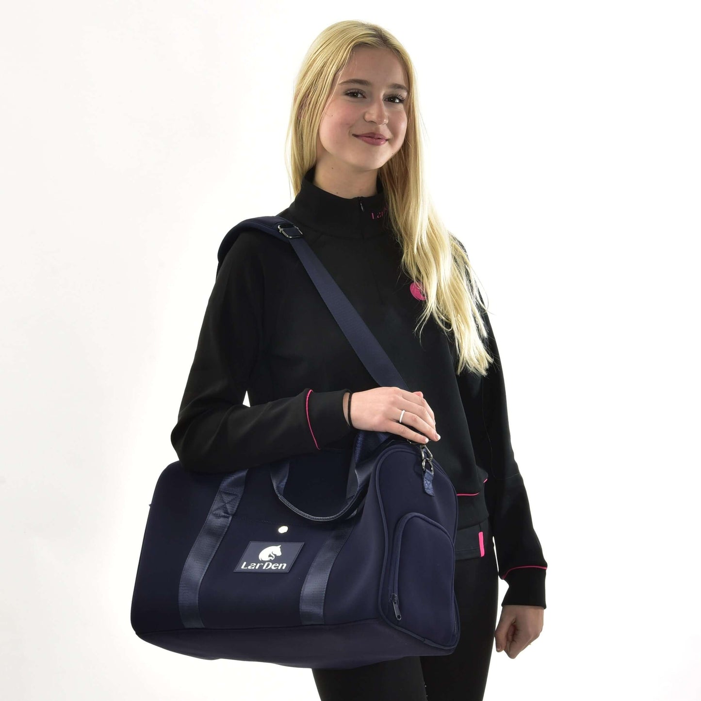 Femme souriante aux longs cheveux blonds, en veste noire à bordure rose, sur fond blanc, tenant le Sac de sport LarDen de Hobby Horse LarDen, sac bleu marine avec sangle réglable, idéal pour un usage quotidien.