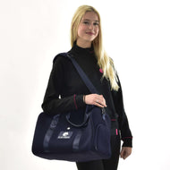 Eine lächelnde Frau mit langen blonden Haaren in einer schwarzen Jacke mit pinken Details steht vor weißem Hintergrund und hält die Sporttasche LarDen von Hobby Horse LarDen – eine dunkelblaue Tasche mit verstellbarem Gurt, ideal für den Alltag.