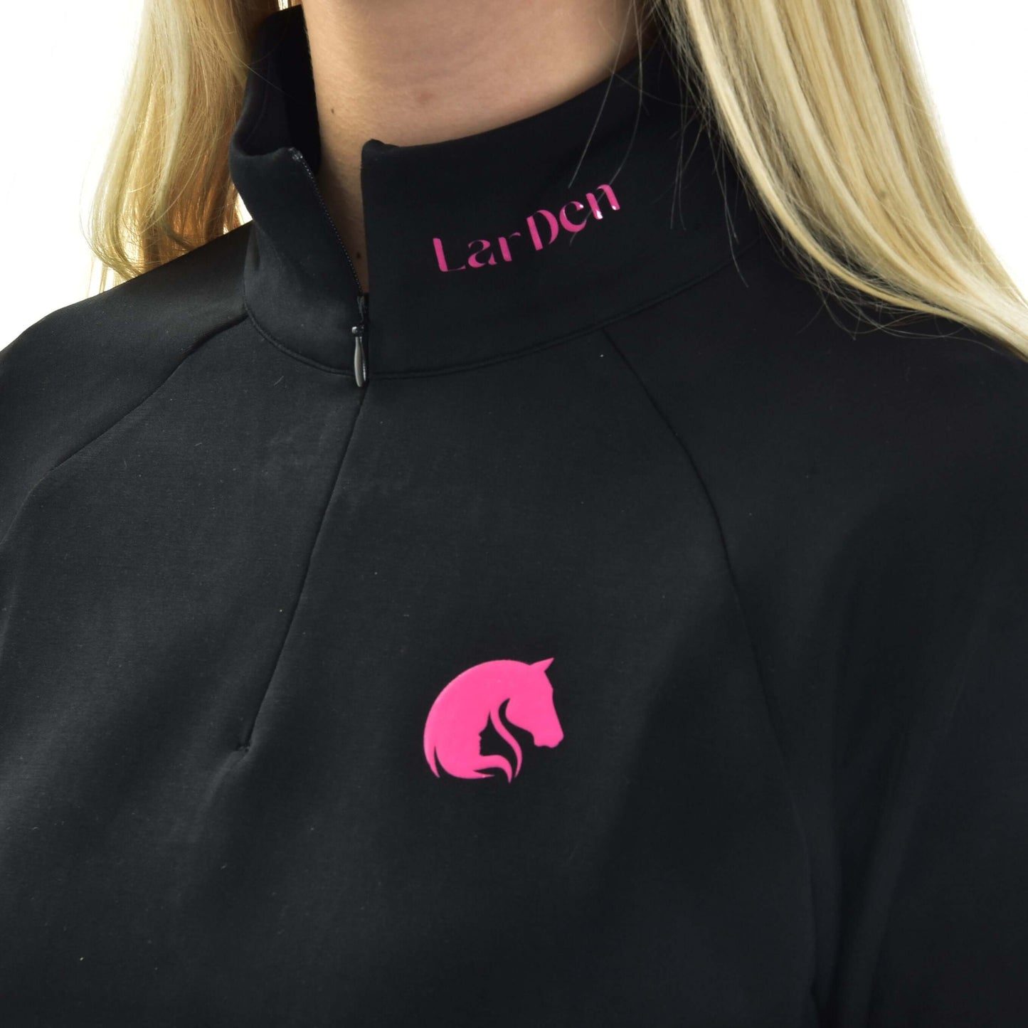 Personne aux longs cheveux blonds portant un sweat-shirt LarDen de Hobby Horse LarDen, un haut noir zippé avec logo tête de cheval rose sur la poitrine et Lar Den en rose près du col.