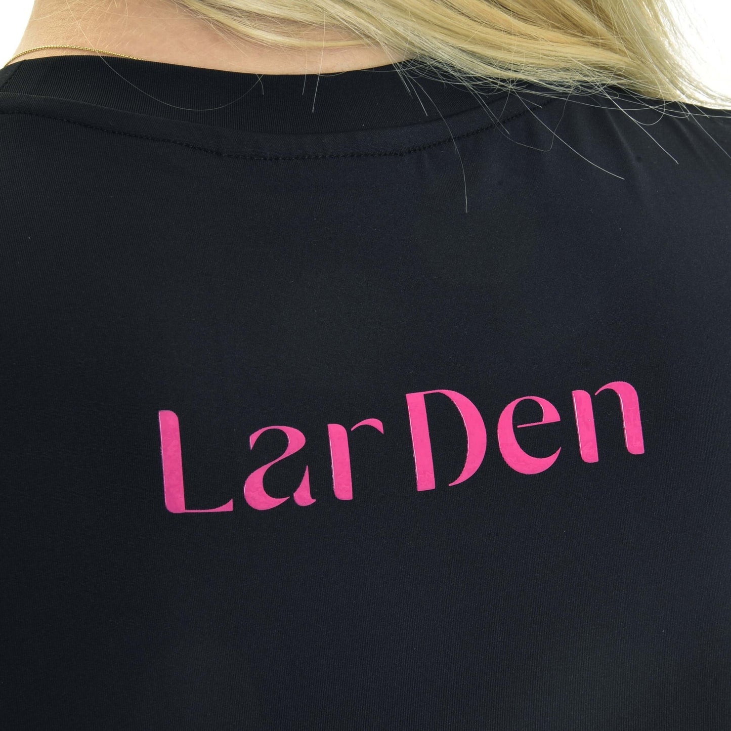 Gros plan du dos d’une personne portant un sweat-shirt LarDen noir de Hobby Horse LarDen avec grand lettrage rose. Des cheveux blonds sont partiellement visibles en haut de l’image.