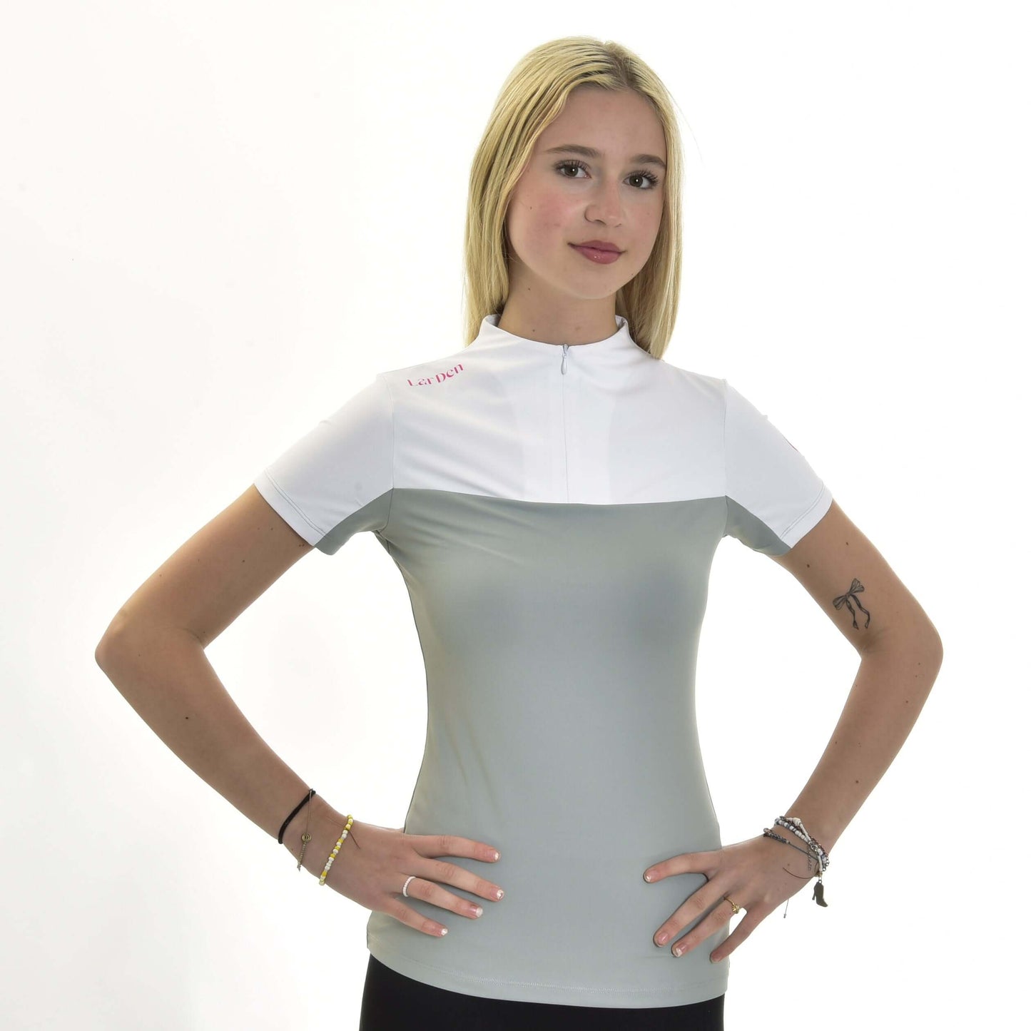 Jeune femme aux longs cheveux blonds portant le T-shirt LarDen Premium de Hobby Horse LarDen, un haut de sport en spandex gris clair et blanc à manches courtes. Elle fait face à l’appareil, mains sur les hanches, avec bracelets et petit tatouage sur le bras gauche visibles.