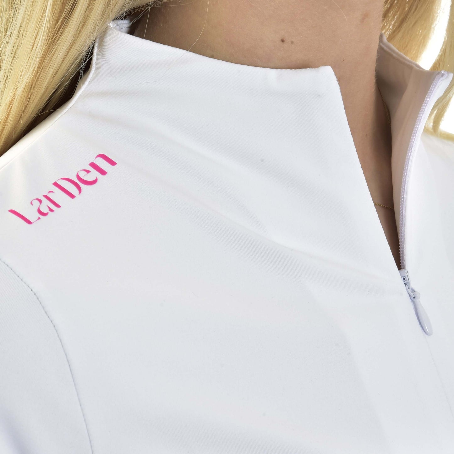 Gros plan d’une personne blonde portant le T-shirt LarDen Premium de Hobby Horse LarDen, t-shirt de sport blanc à fermeture zippée avec inscription rose « La Den » sur l’épaule. Le tissu en spandex assure le confort.