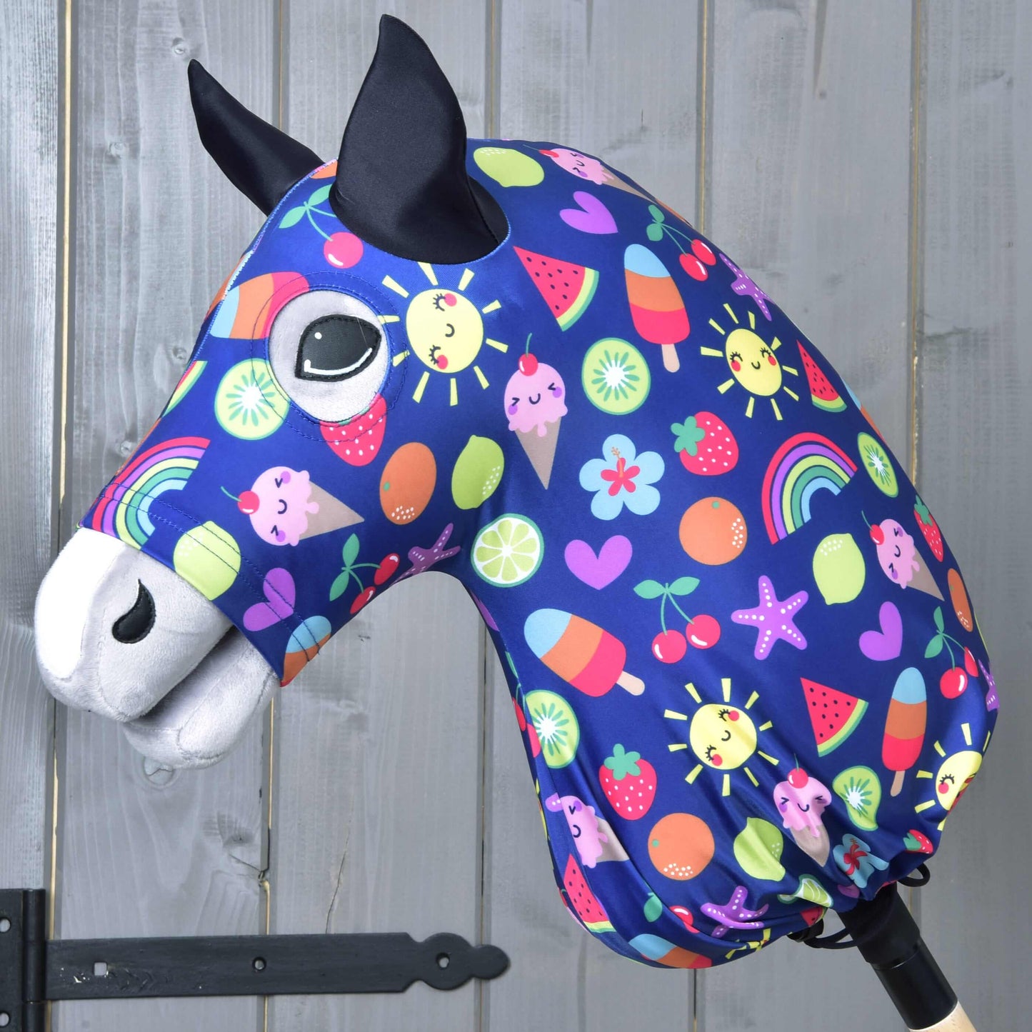 Tête de Hobby Horse en tissu avec oreilles noires, grands yeux et combinaison de protection Hobby Horse Bonheur de Hobby Horse LarDen bleue avec soleils, arcs-en-ciel, cœurs, fruits et glaces, sur fond en bois.