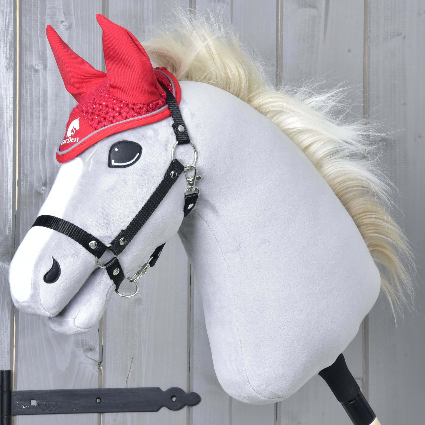 Ein plüschiges Hobby Horse von Hobby Horse LarDen mit weißem Gesicht, schwarzem Zaumzeug und blonder Mähne präsentiert die Ohrenkappe gehäkelt Red Sport vor einer hellen Holzwand.