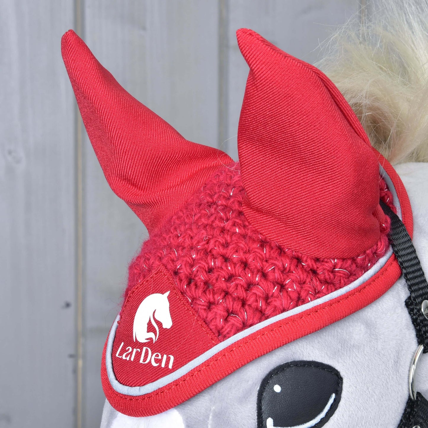 Nahaufnahme eines Pferdes mit der Hobby Horse LarDen Ohrenkappe gehäkelt Red Sport mit weißem LarDen-Logo und Pferdegrafik, vor hellgrauen Holzpaneelen.