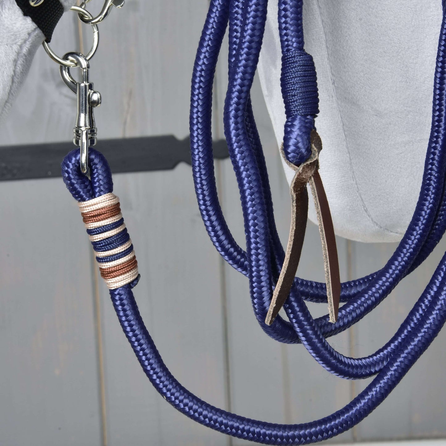 Hobby Horse - Longe / Longierseil, Nahaufnahme einer blauen geflochtenen Longe von Hobby Horse LarDen mit silberfarbenem Karabiner und Lederquaste, mit beigem und schwarzem Zierfaden nahe dem Karabiner, hängend vor Holz-Hintergrund.