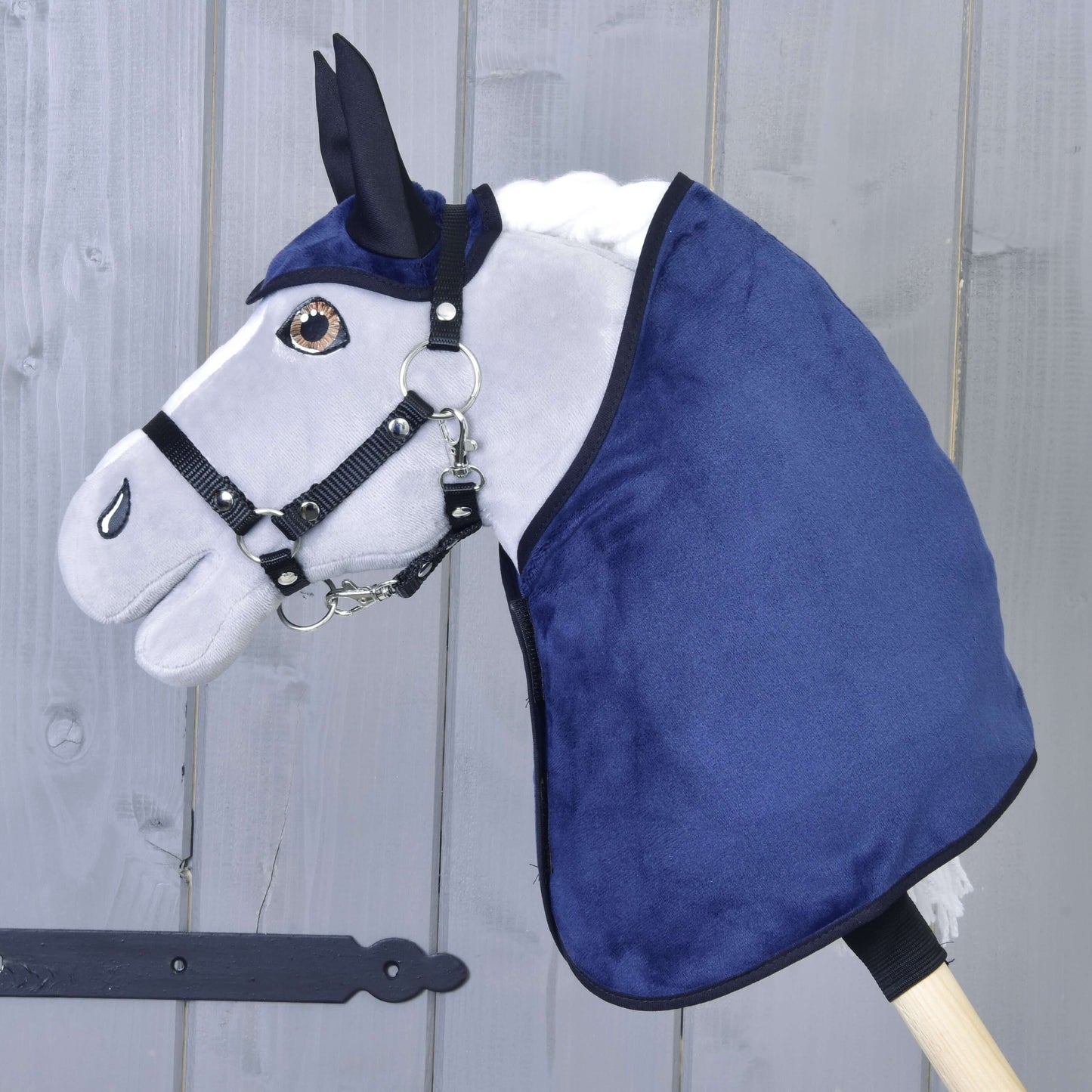 Hobby Horse LarDen Set Dark Blue (Ohrenkappe + Decke) mit plüschigem Hobby Horse-Kopf, weißer Mähne, schwarzem Zaumzeug, blauer Ohrenkappe und Decke, auf einem Holzstab vor einem hellen Holzzaun.