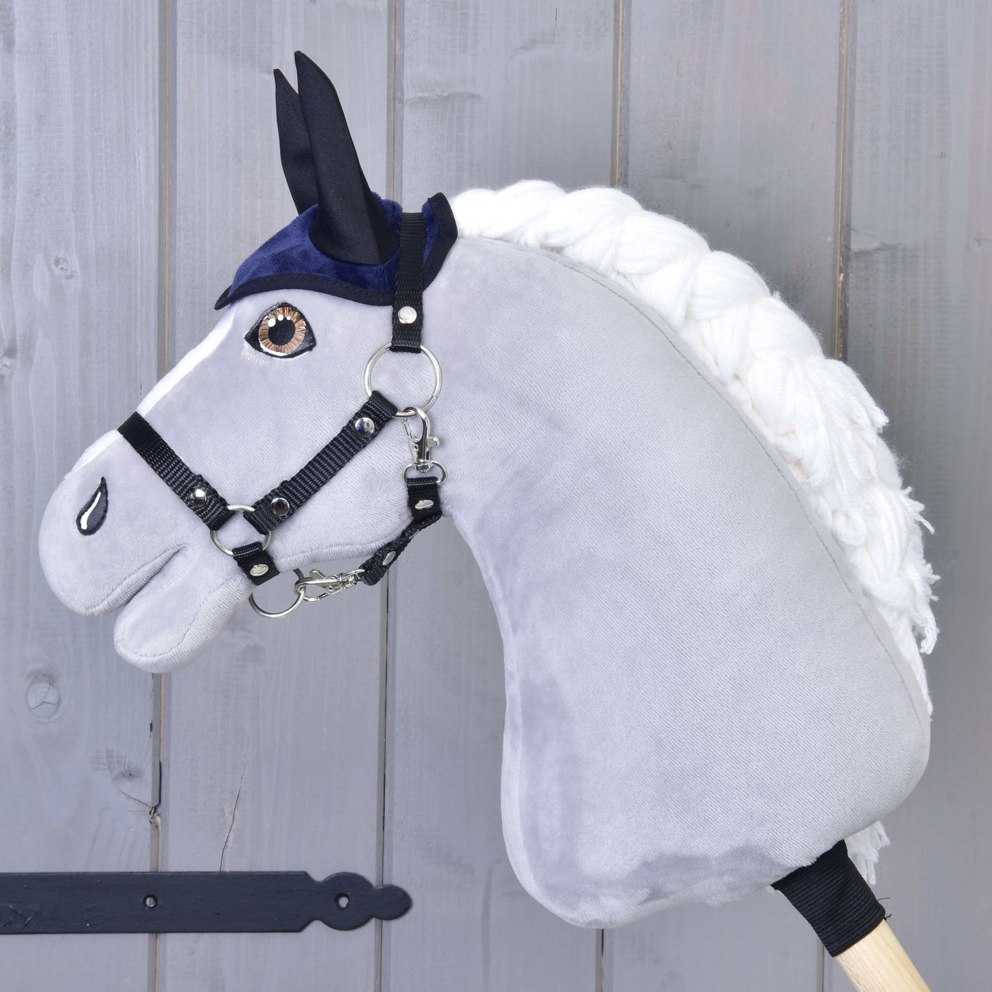 Plüschiges Hobby Horse von Hobby Horse LarDen mit weißer Mähne, schwarzem Zaumzeug und dem Set Dark Blue (Ohrenkappe + Decke), auf einem Holzstab vor einer hellgrauen Holzwand.