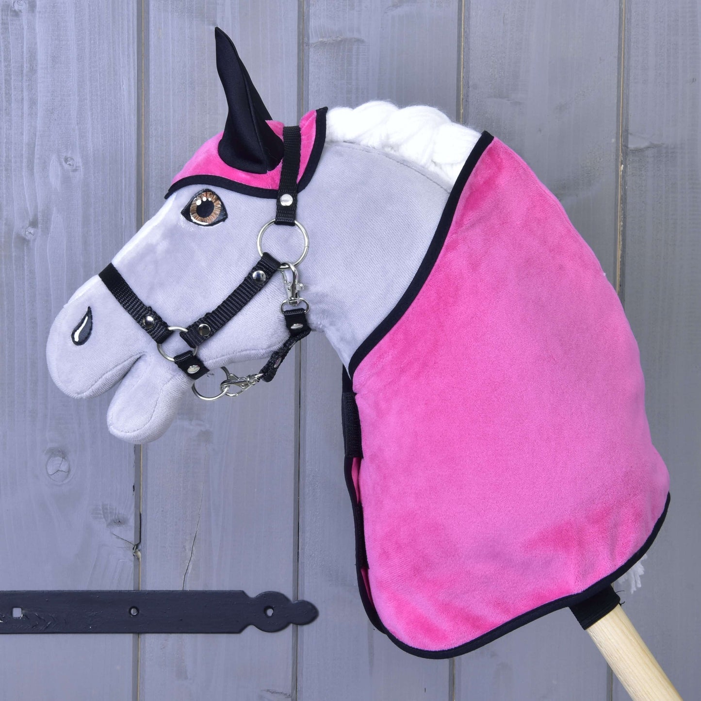 Graues Steckenpferd von Hobby Horse LarDen mit Set Hot Pink (Ohrenkappe + Decke) und schwarzem Zaumzeug vor einem Holzzaun.