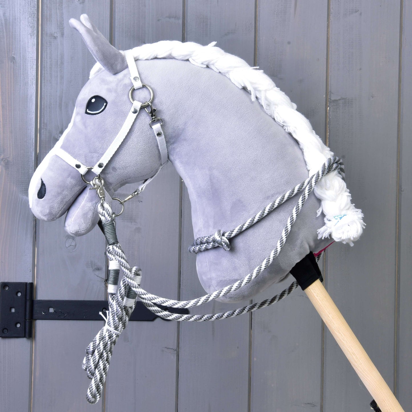 Ein graues Plüsch-Hobby Horse von Hobby Horse LarDen mit Set Start Silber (Halfter + Halsring + Führstrick + Zügel), an einem Holzstab befestigt und vor Holzpaneel-Hintergrund gezeigt.