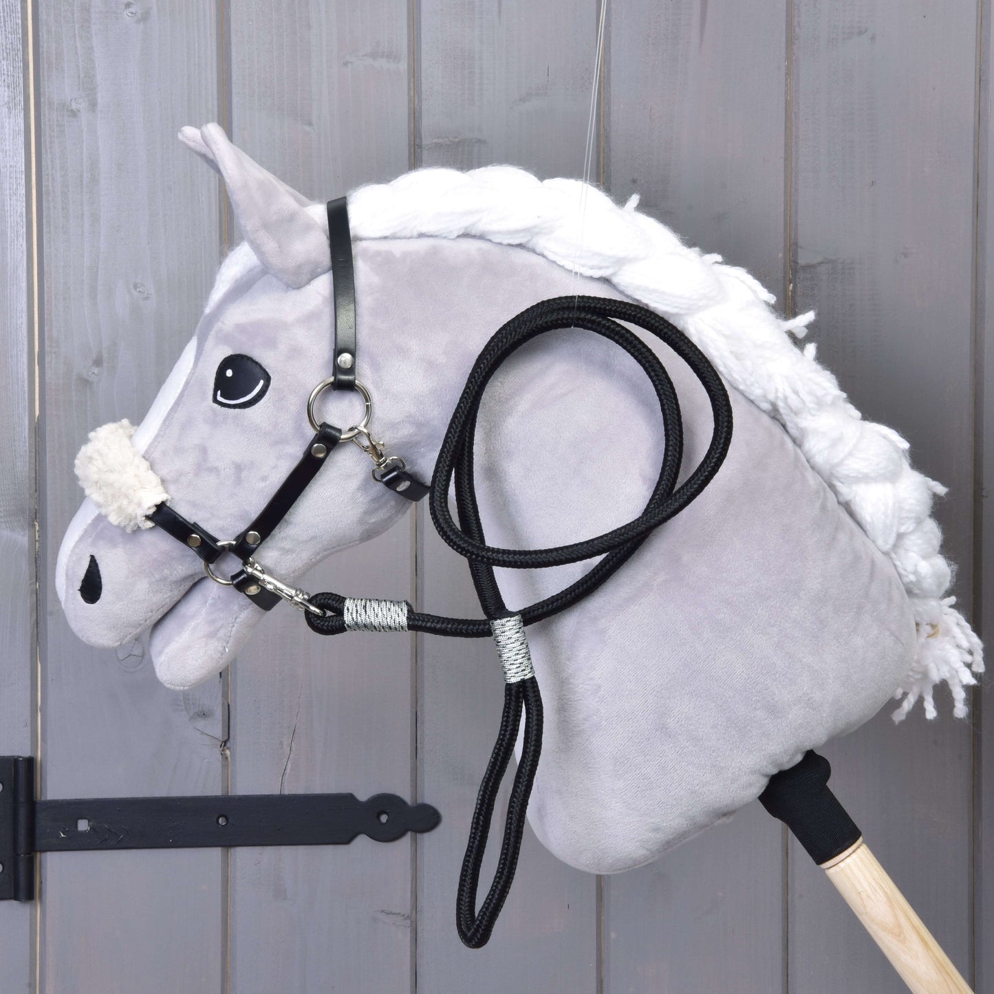 Das Hobby Horse LarDen Set Basic zeigt ein Plüsch-Hobby Horse mit weißer Mähne, schwarzem Zaumzeug mit Zügeln und schwarzem Halfter für Hobby Horse auf einem Holzstab, angelehnt an graue Holzpaneele.