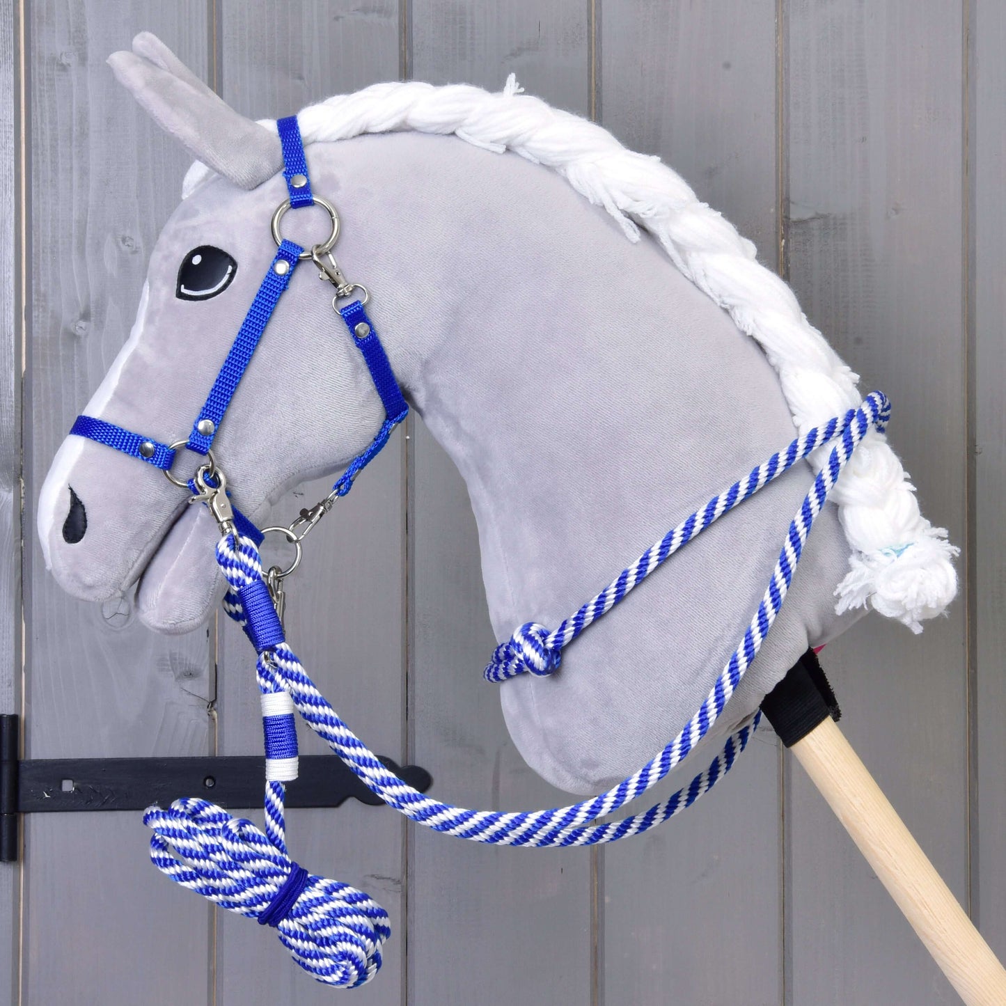 Ein graues Plüsch-Hobby Horse von Hobby Horse LarDen mit Set Start Blau (Halter, Halsring, Führstrick, Zügel), mit blau-weißem Führstrick an einem Holzstab, steht vor einer grauen Holzwand.