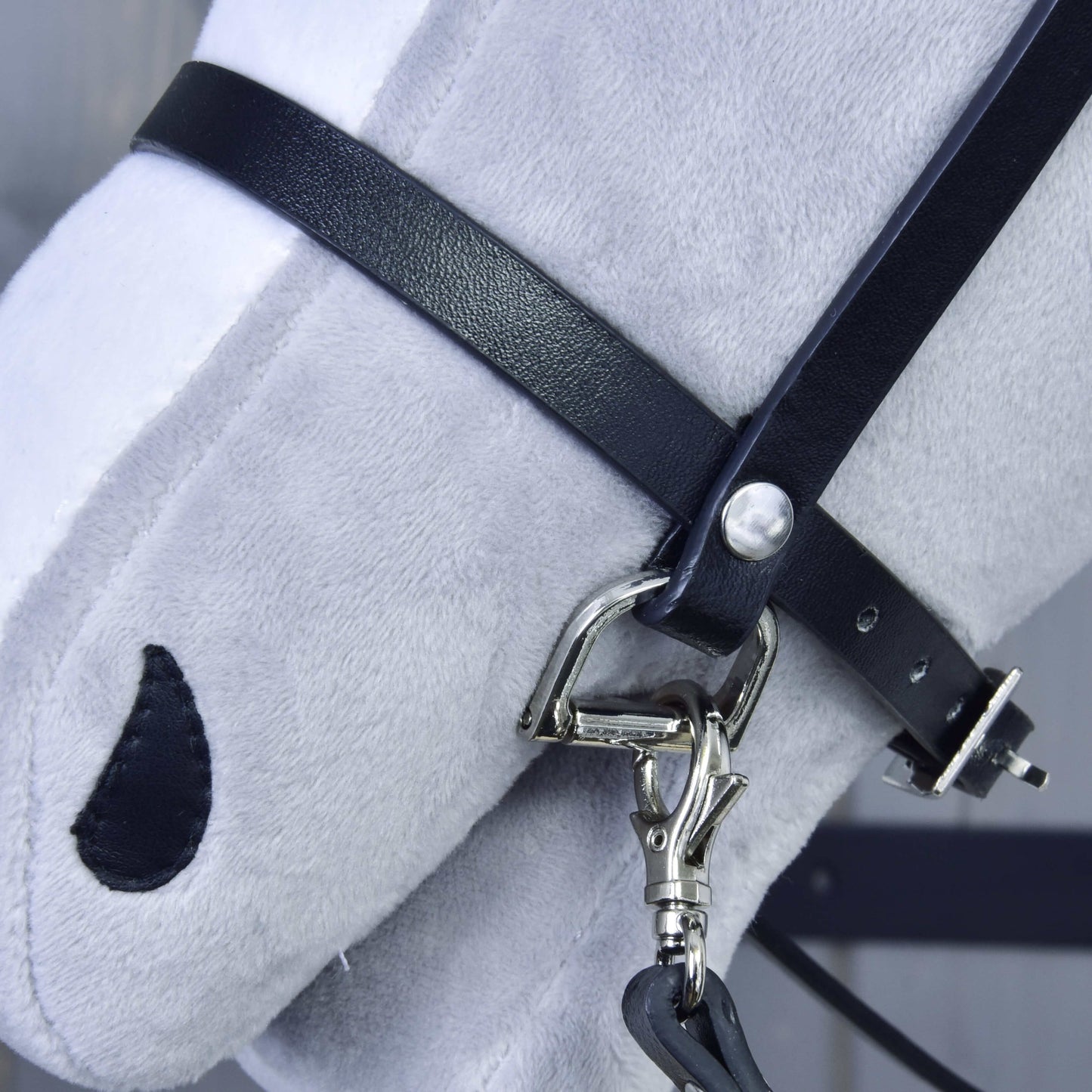 Nahaufnahme des Kopfes eines hellgrauen Plüschpferds mit Schwarzes Zaumzeug aus Kunstleder von Hobby Horse LarDen, mit Metallbeschlägen und einem einfachen schwarz gestickten Auge.